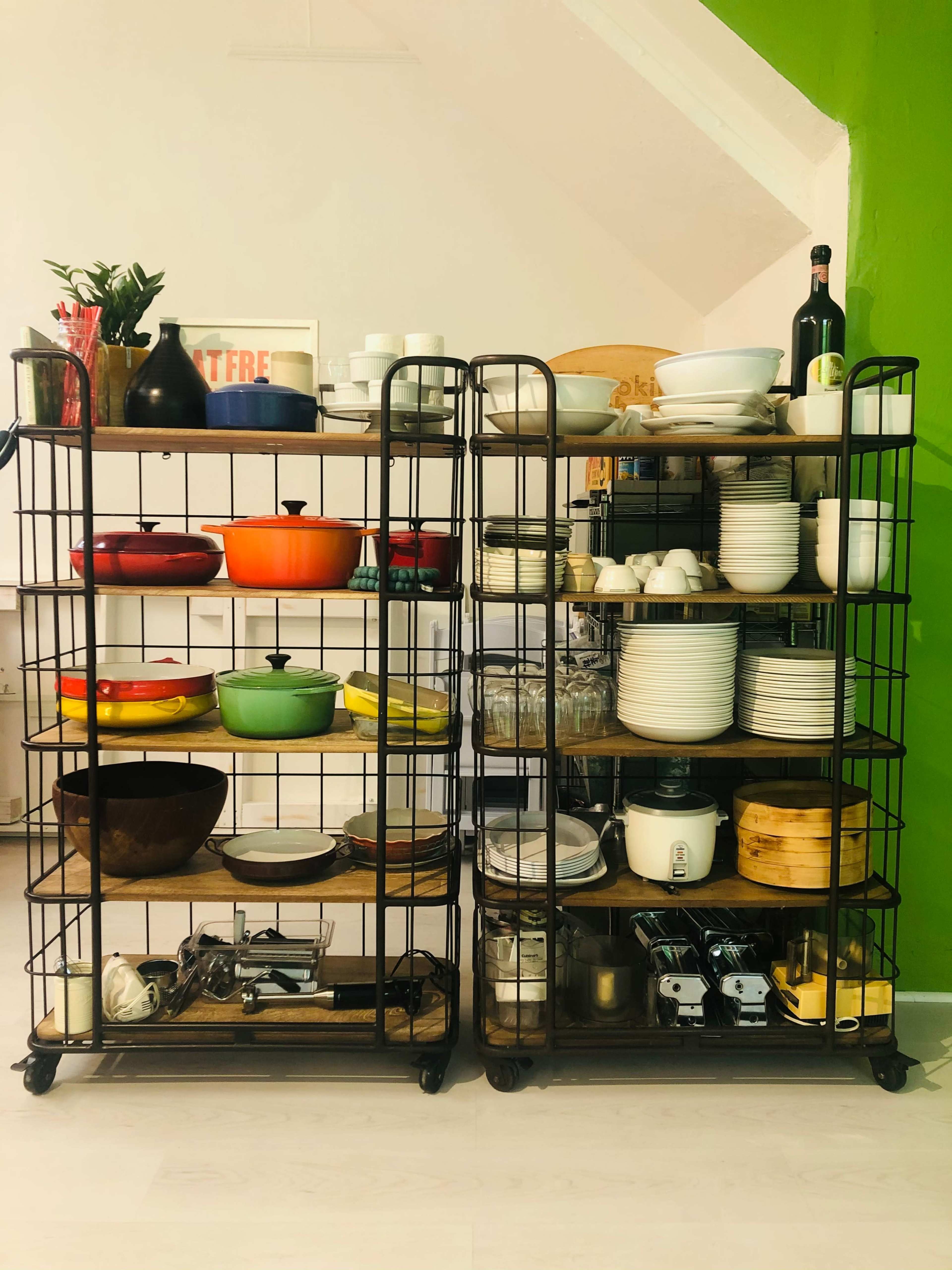 Two metal shelving units filled with various kitchenware, including colorful bowls and white dishes, are positioned against a green wall.