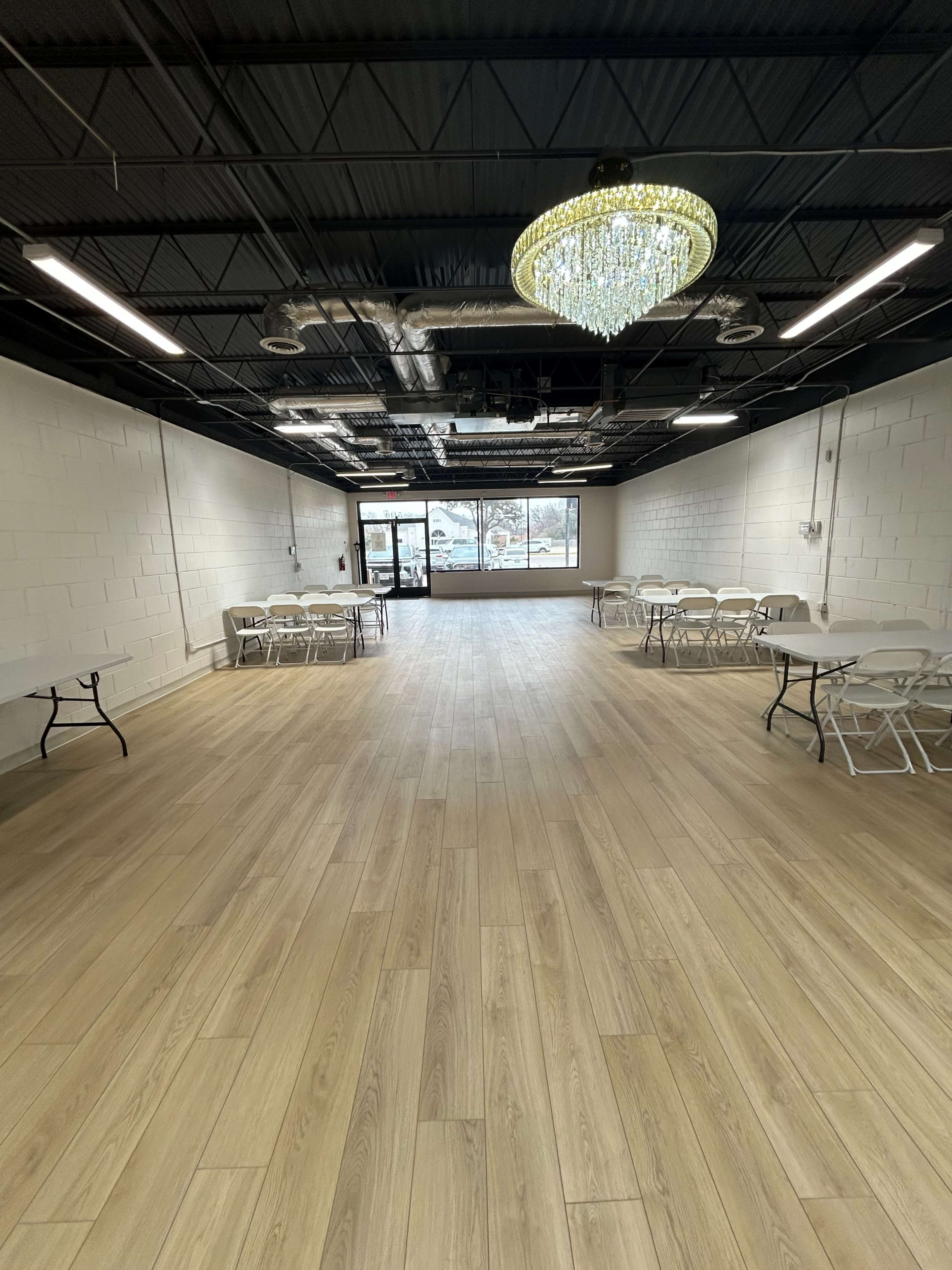 The image shows a spacious indoor venue with wooden flooring, several round tables, and a large window at one end, illuminated by a chandelier.