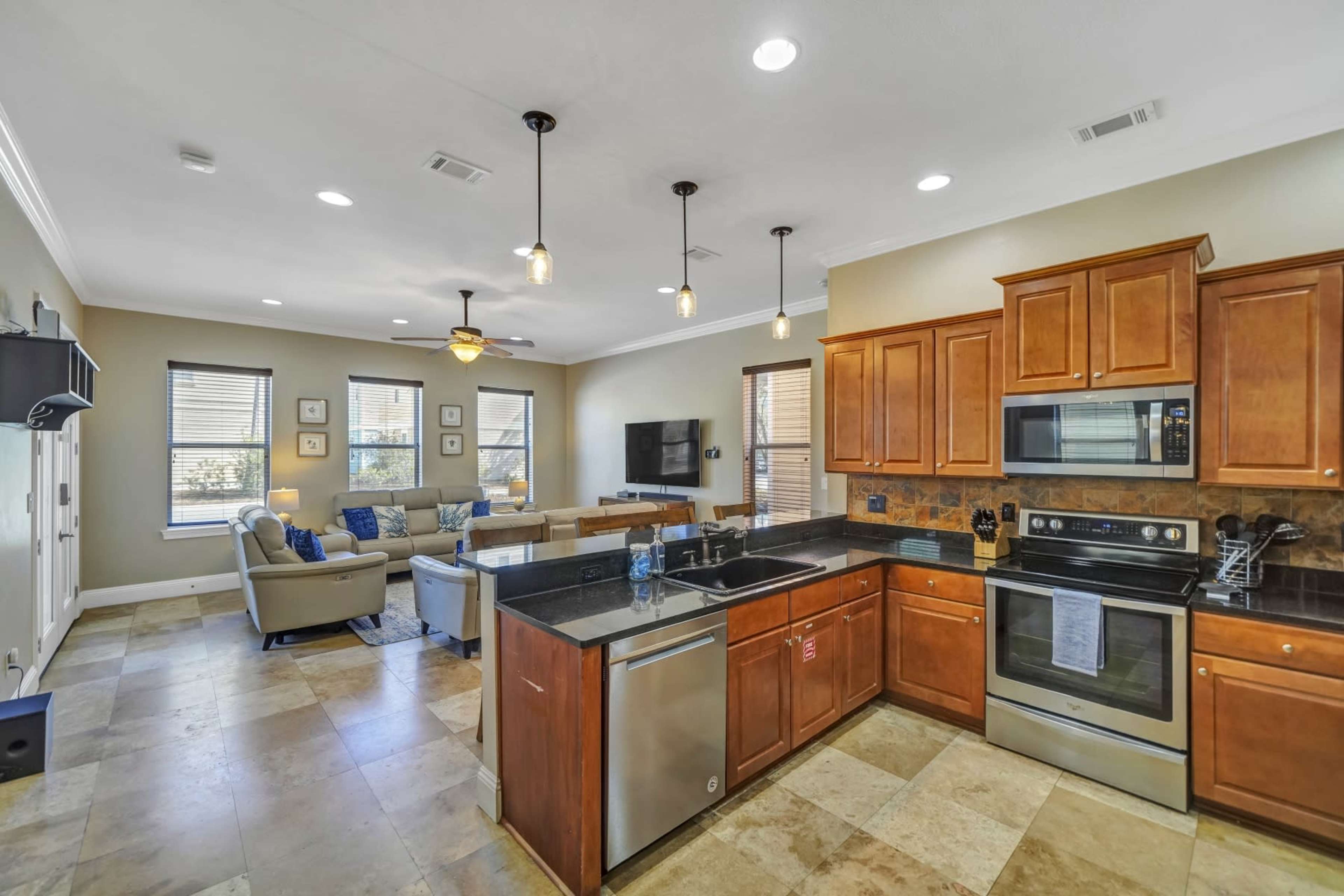 A modern kitchen with wooden cabinets and stainless steel appliances, opening into a living area with sofas and a television.