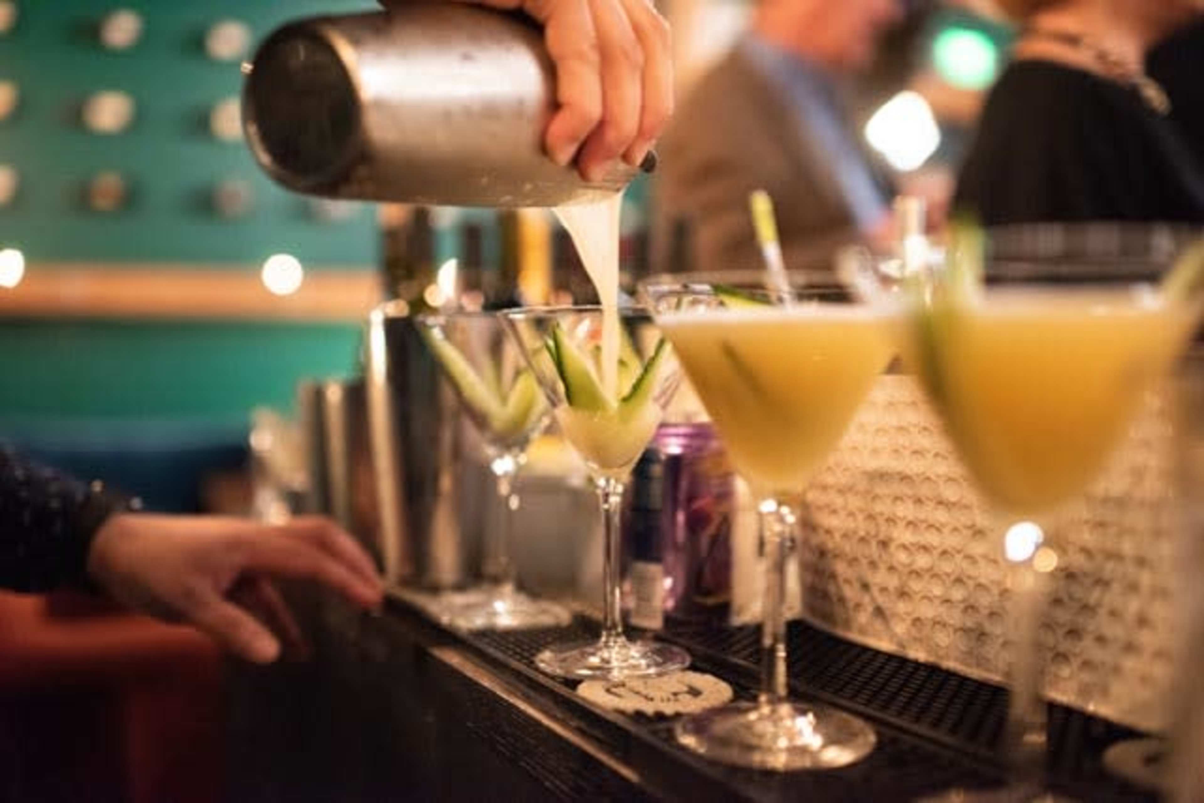 A bartender pours a yellow cocktail from a shaker into a martini glass garnished with cucumber at a bar.