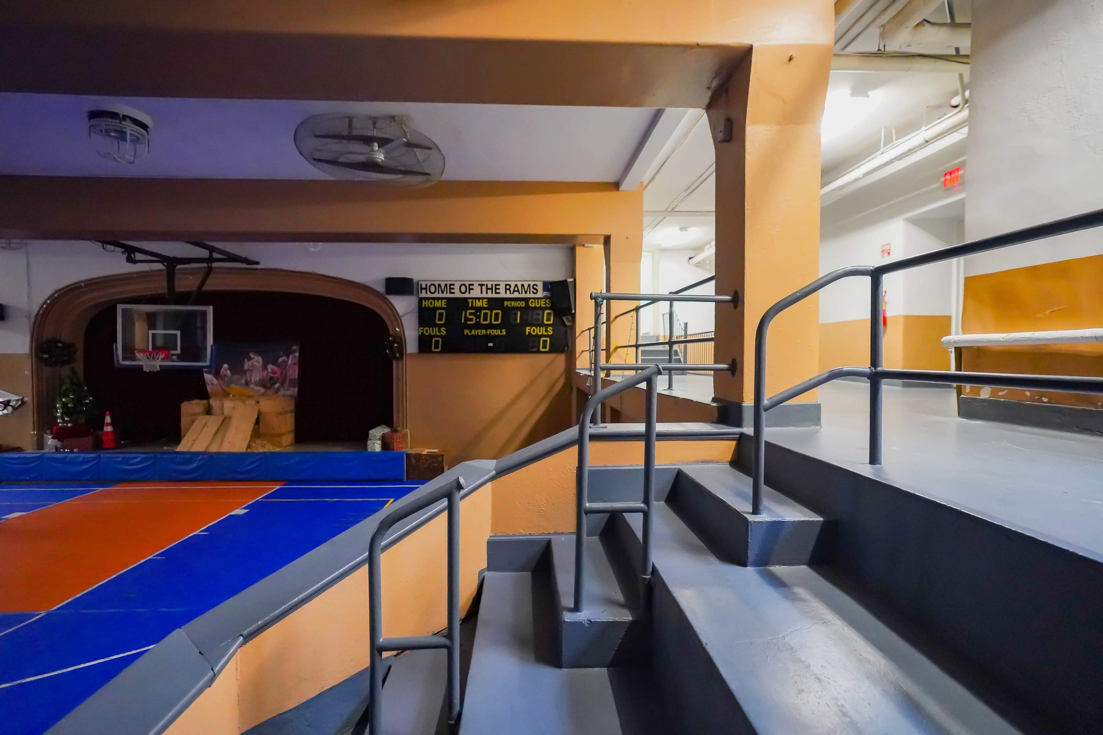 A gymnasium interior with a basketball court, scoreboard, and a set of stairs leading to an upper level.