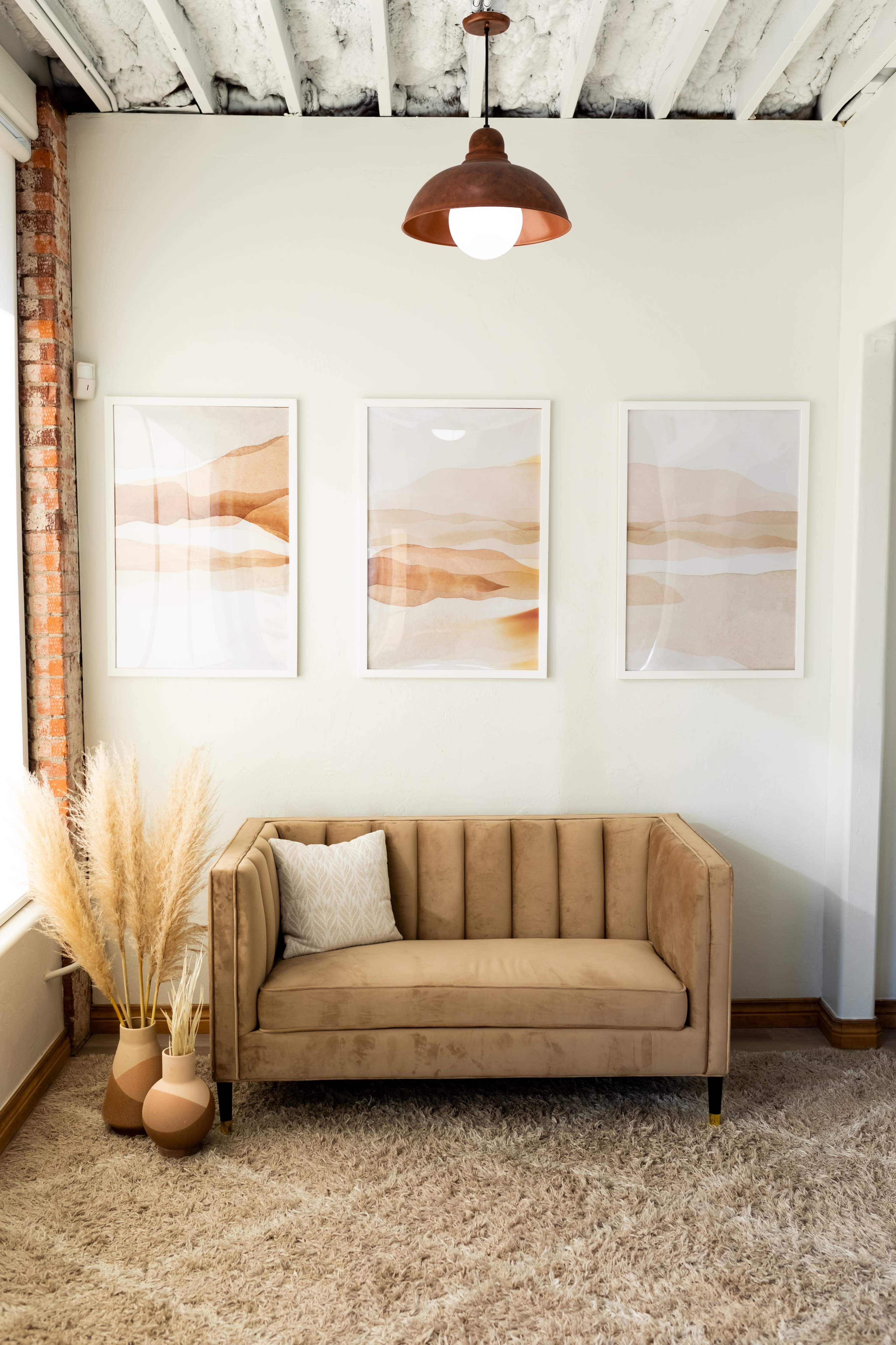 A cozy living room with a beige sofa, three framed art pieces on the wall, and decorative plants in neutral-colored vases.