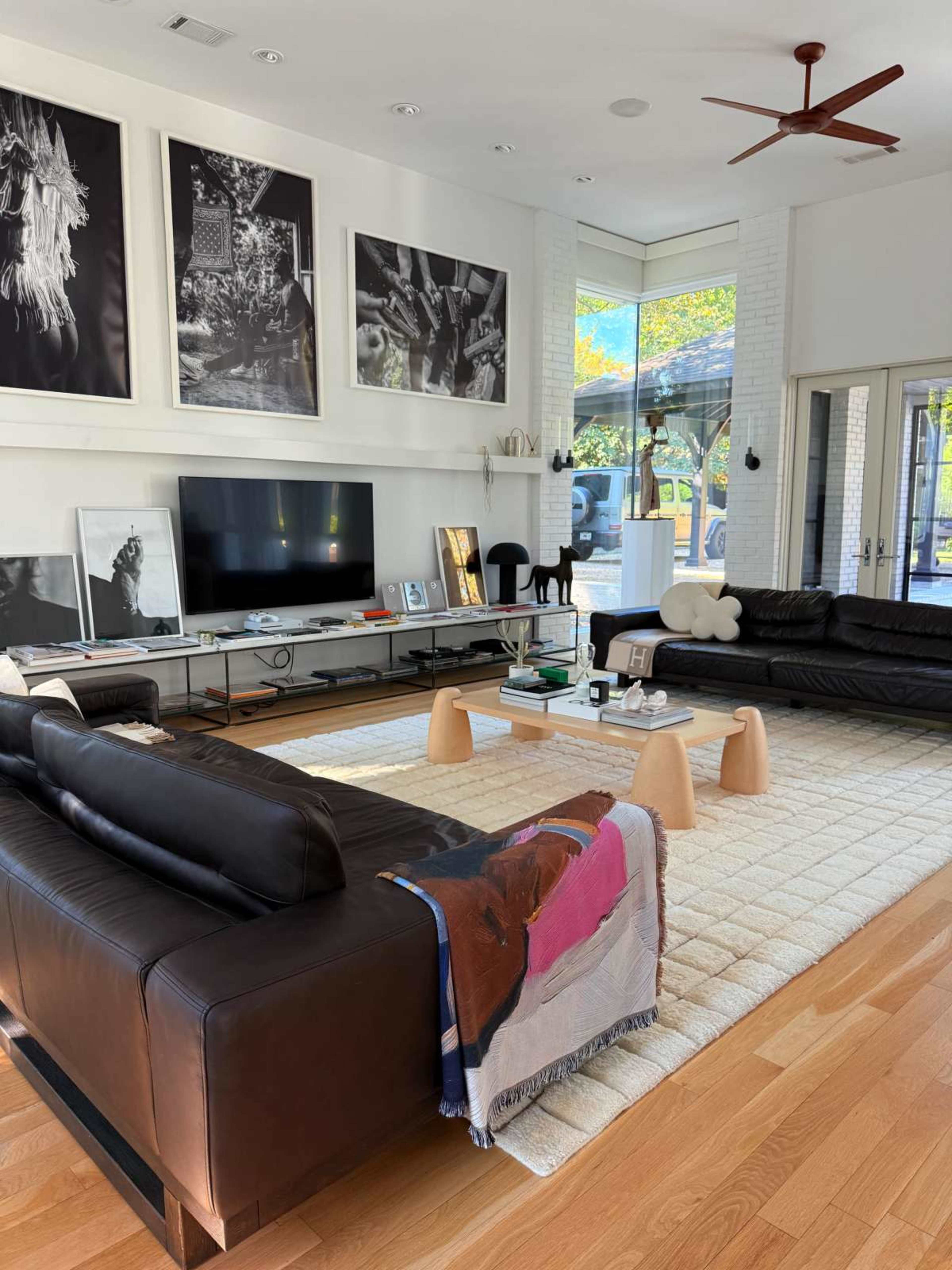 The living room features two dark leather sofas facing each other, a large television mounted on the wall, and a coffee table surrounded by light-colored stools on a textured rug.