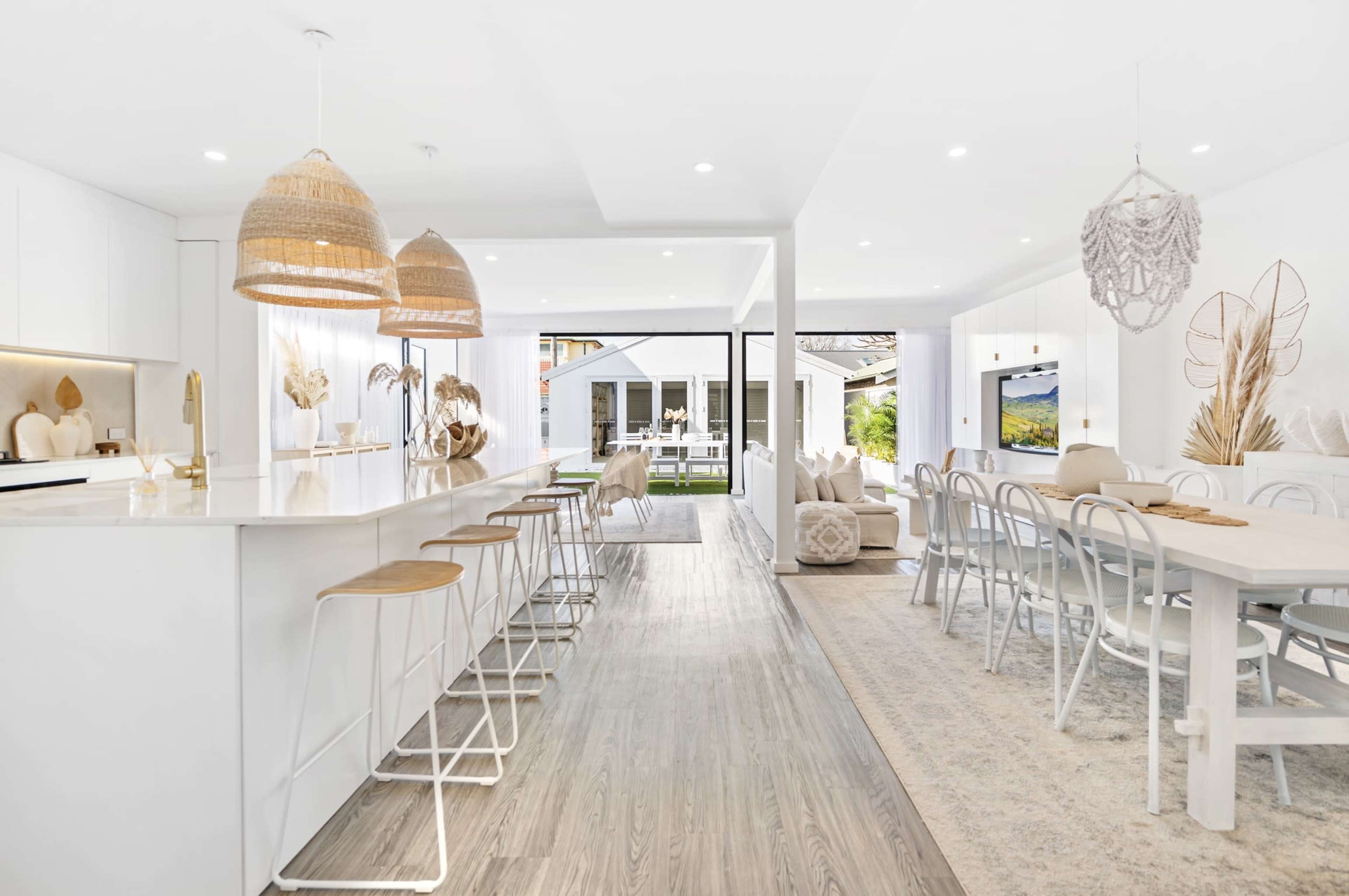 A modern, open-concept kitchen and dining area with white cabinetry, bar stools, and large windows overlooking an outdoor space.