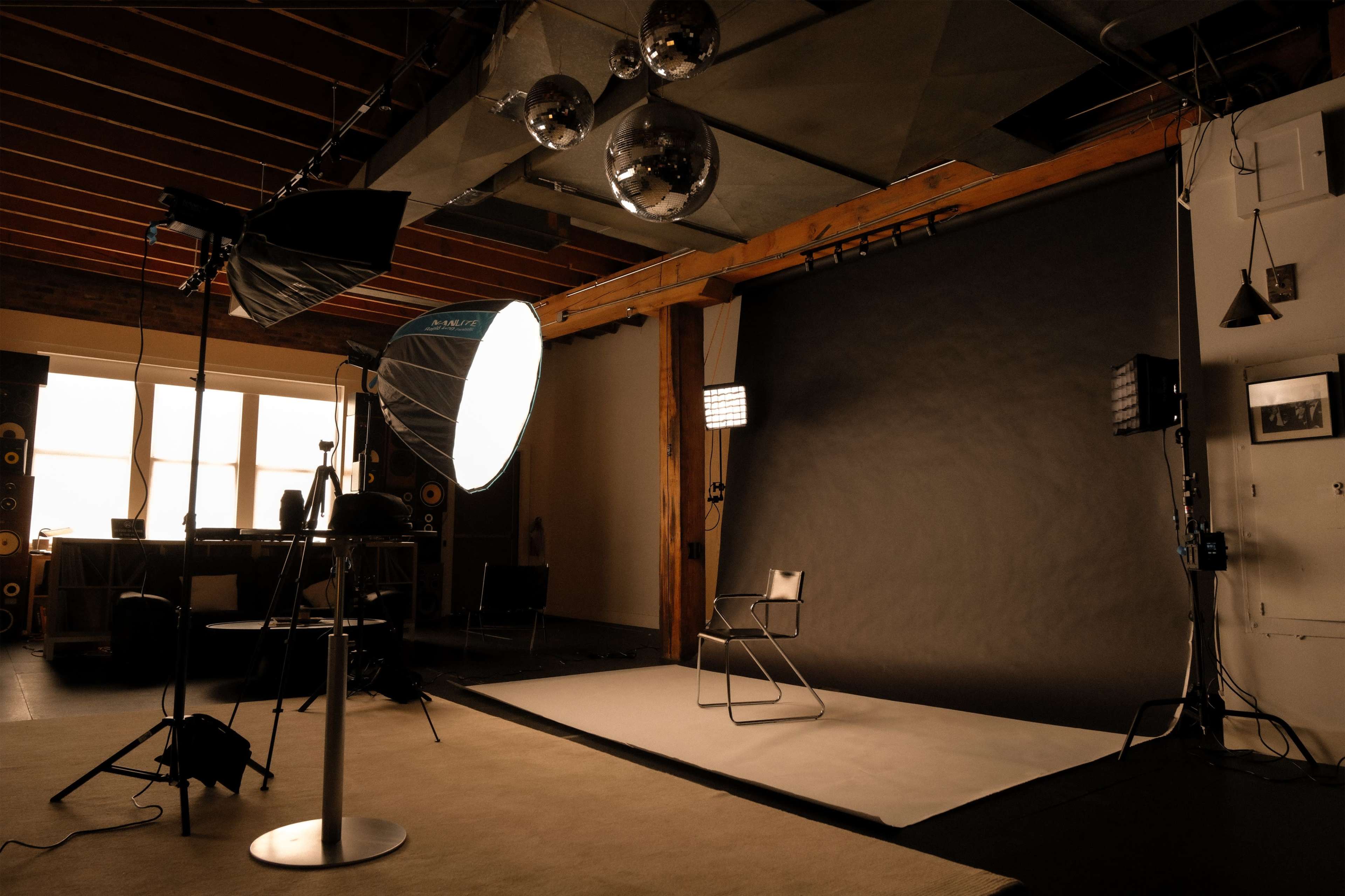 The image shows a photography studio equipped with lighting equipment, a backdrop, and a chair positioned on a neutral-colored surface.