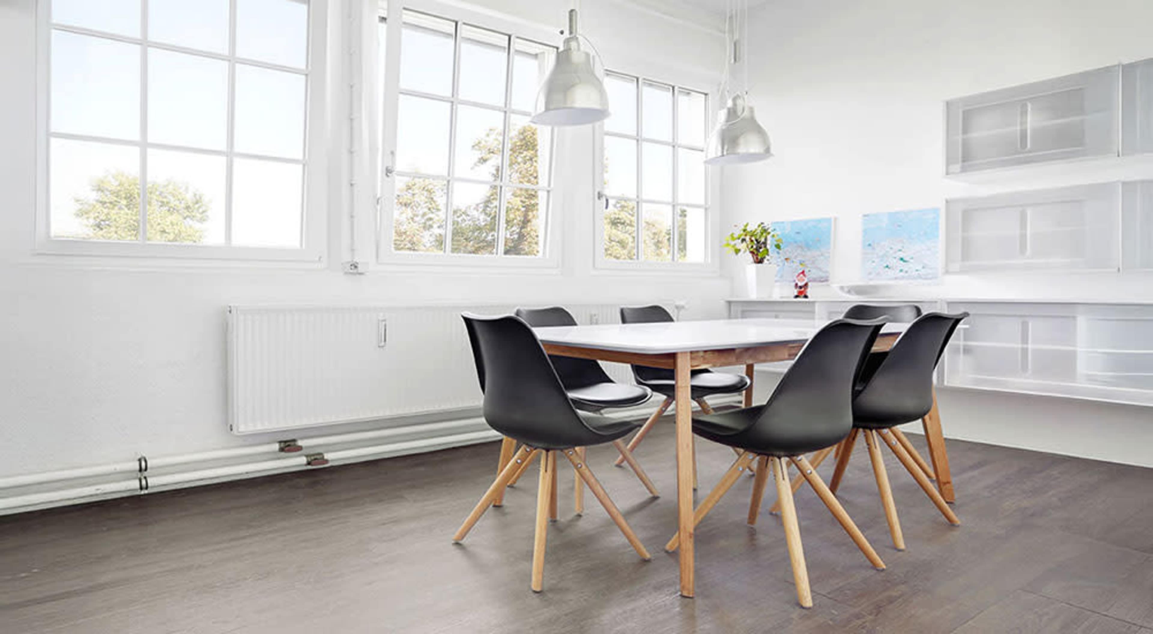 The image shows a minimalist dining area featuring a wooden table surrounded by black chairs, with large windows letting in natural light.