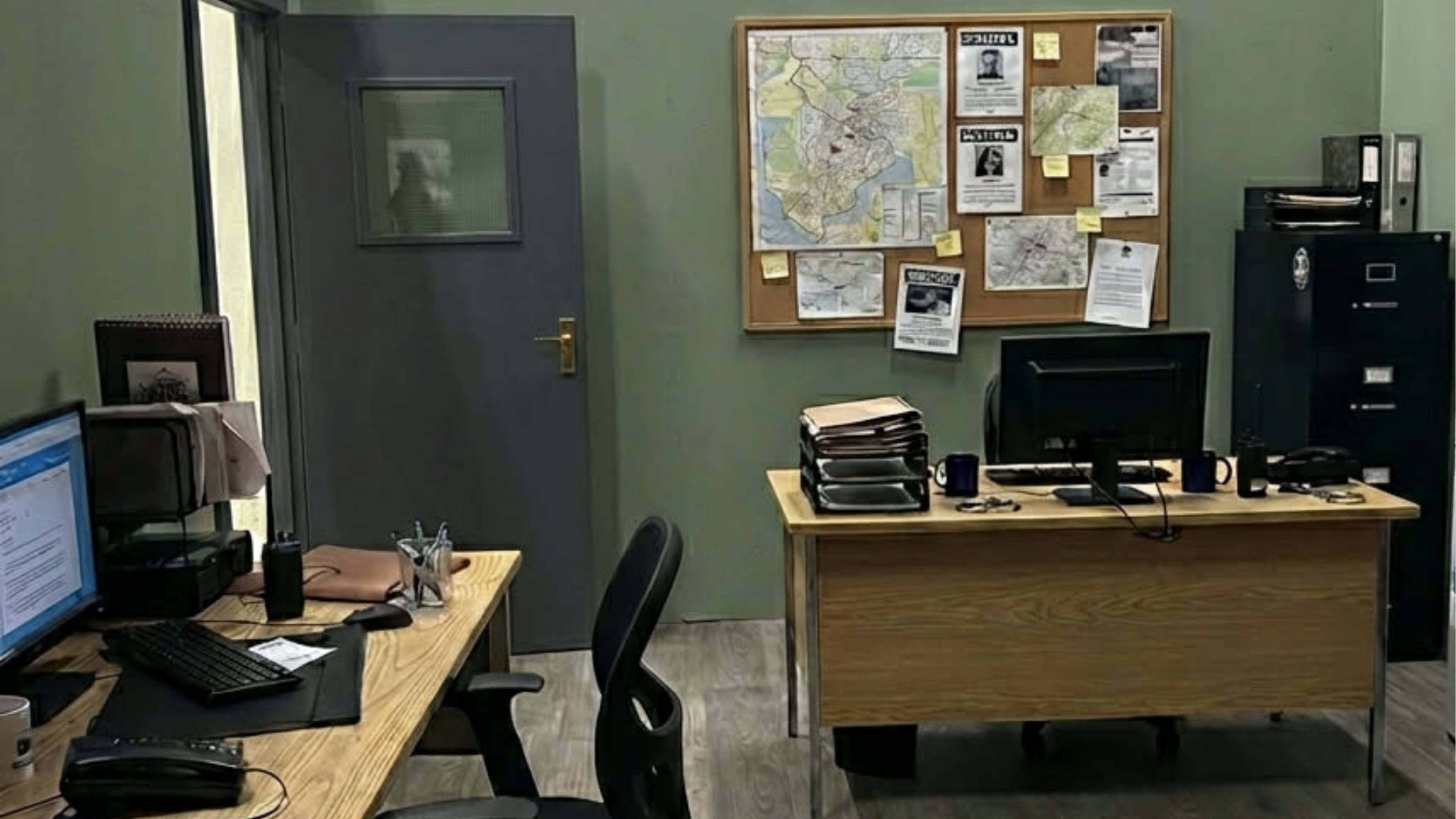 The image depicts a small office setup with two desks, a computer on each, a bulletin board with maps and notices, and a filing cabinet against the wall.
