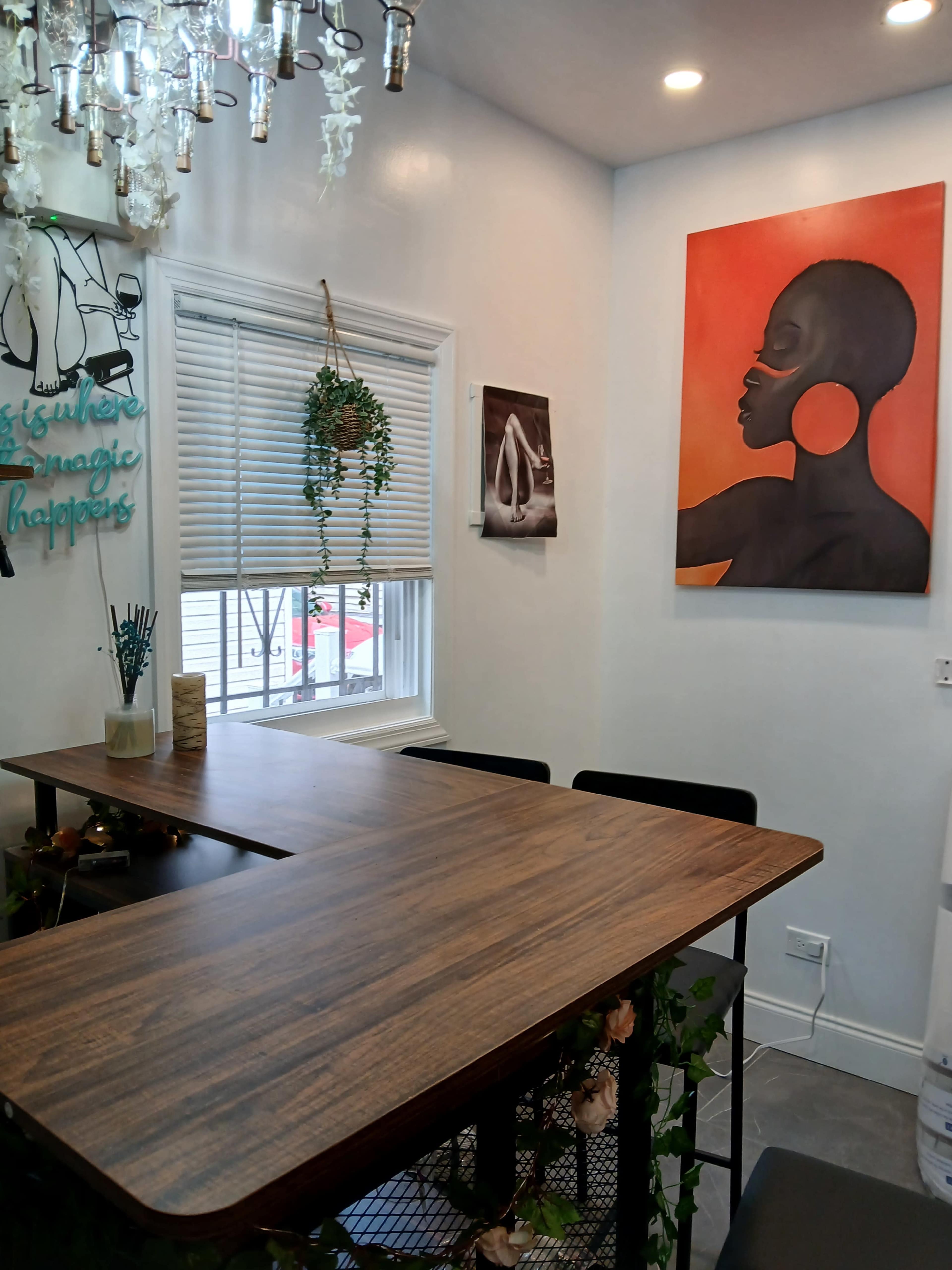 A wooden table with black chairs is positioned in a brightly lit room featuring two wall art pieces and a hanging plant near the window.