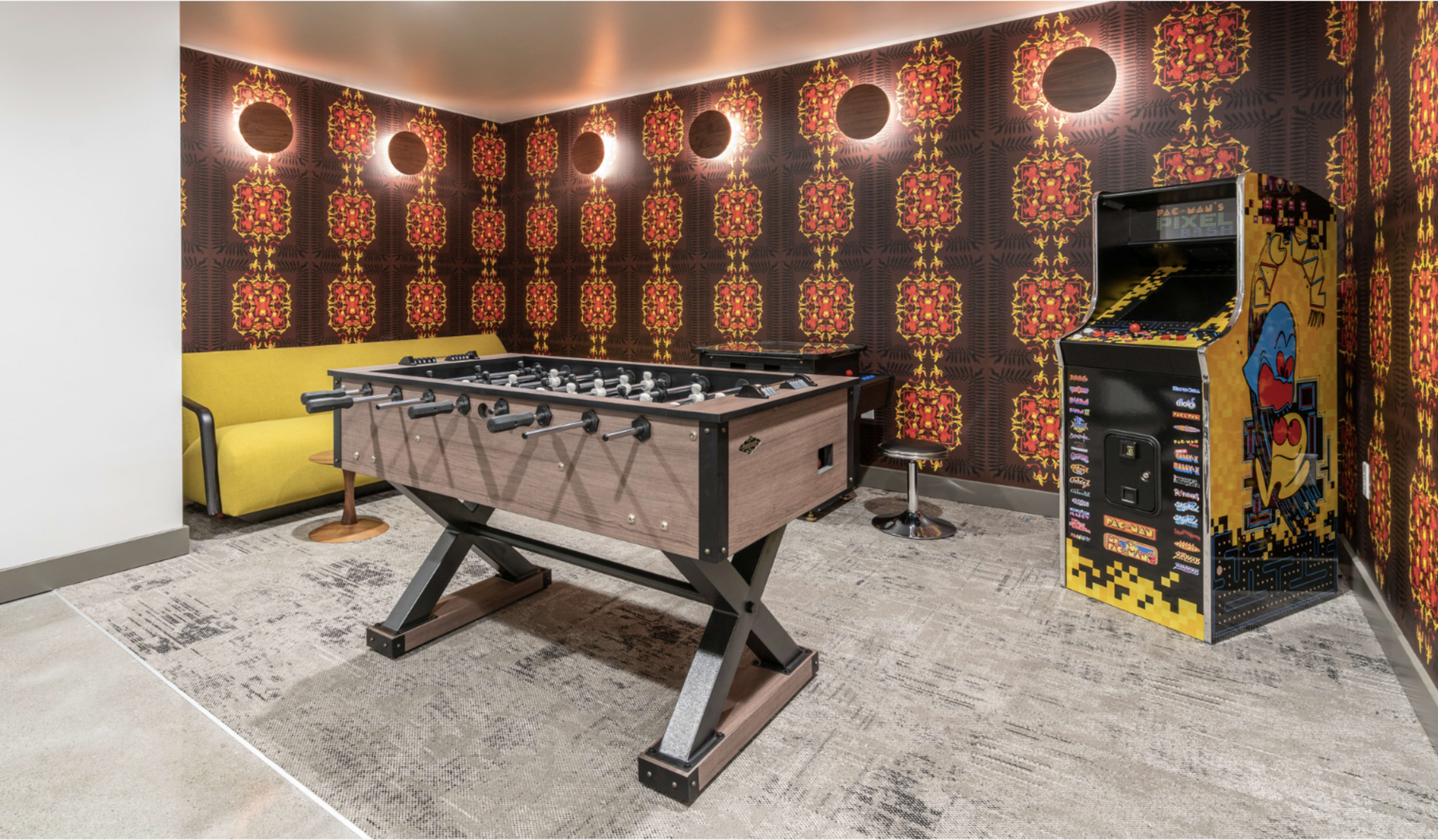 The image shows a game room featuring a foosball table, a vintage arcade machine, a yellow sofa, and patterned wallpaper.