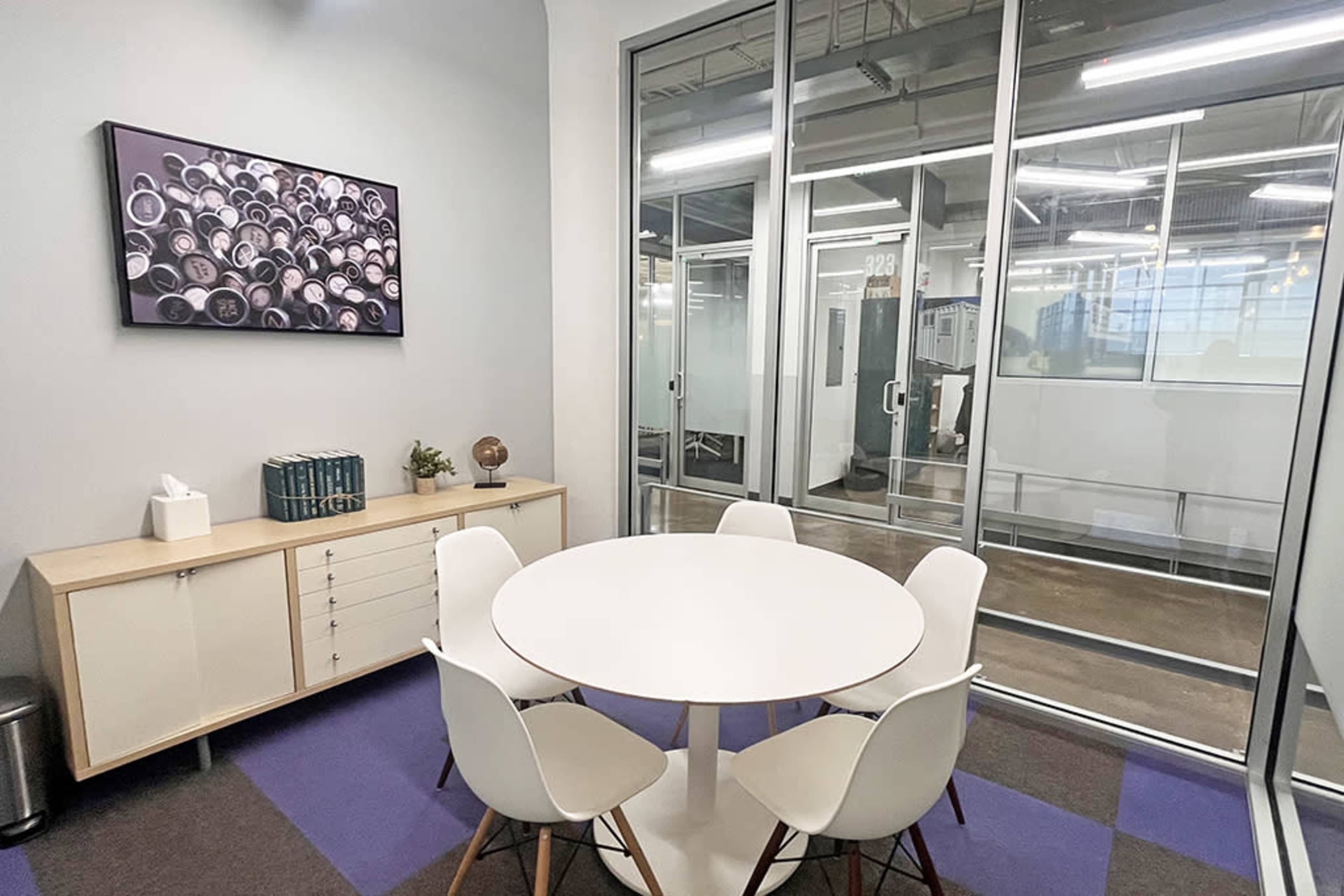The image shows a small meeting room with a round white table surrounded by four chairs, a cabinet, and a wall adorned with a circular art piece.