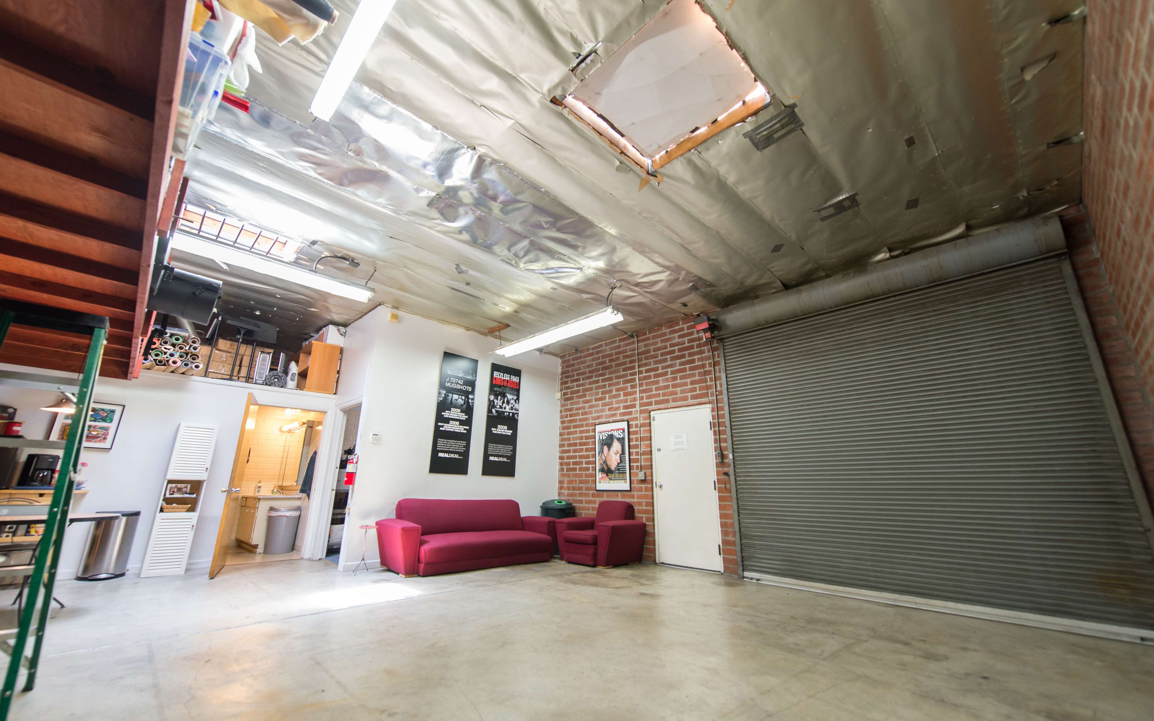 The image shows an industrial-style space with exposed brick walls, a metal rolling door, and a skylight, featuring a red couch and various posters on the wall.