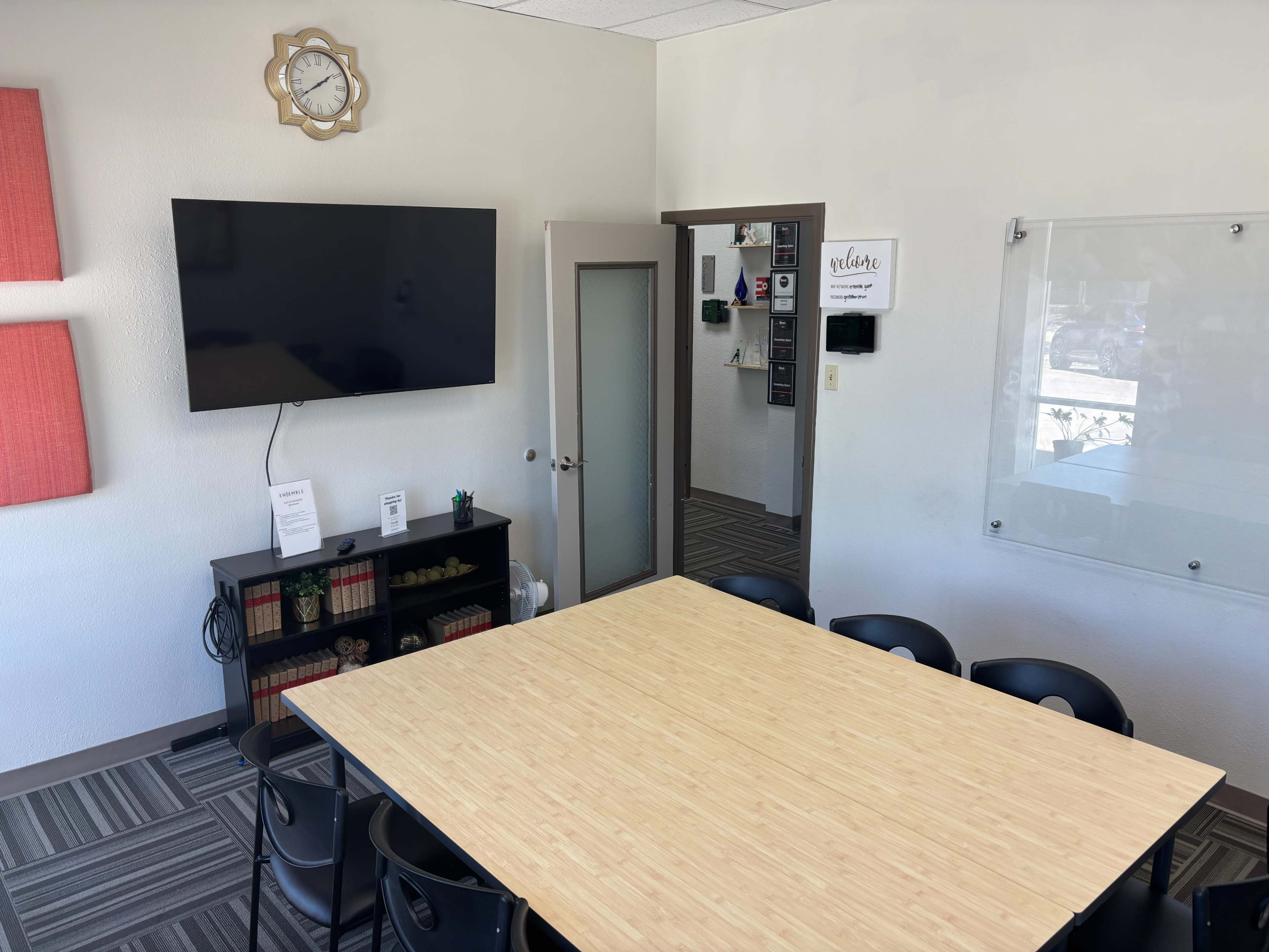 The image shows a meeting room with a large wooden table surrounded by black chairs, a television mounted on the wall, and a whiteboard next to the door.