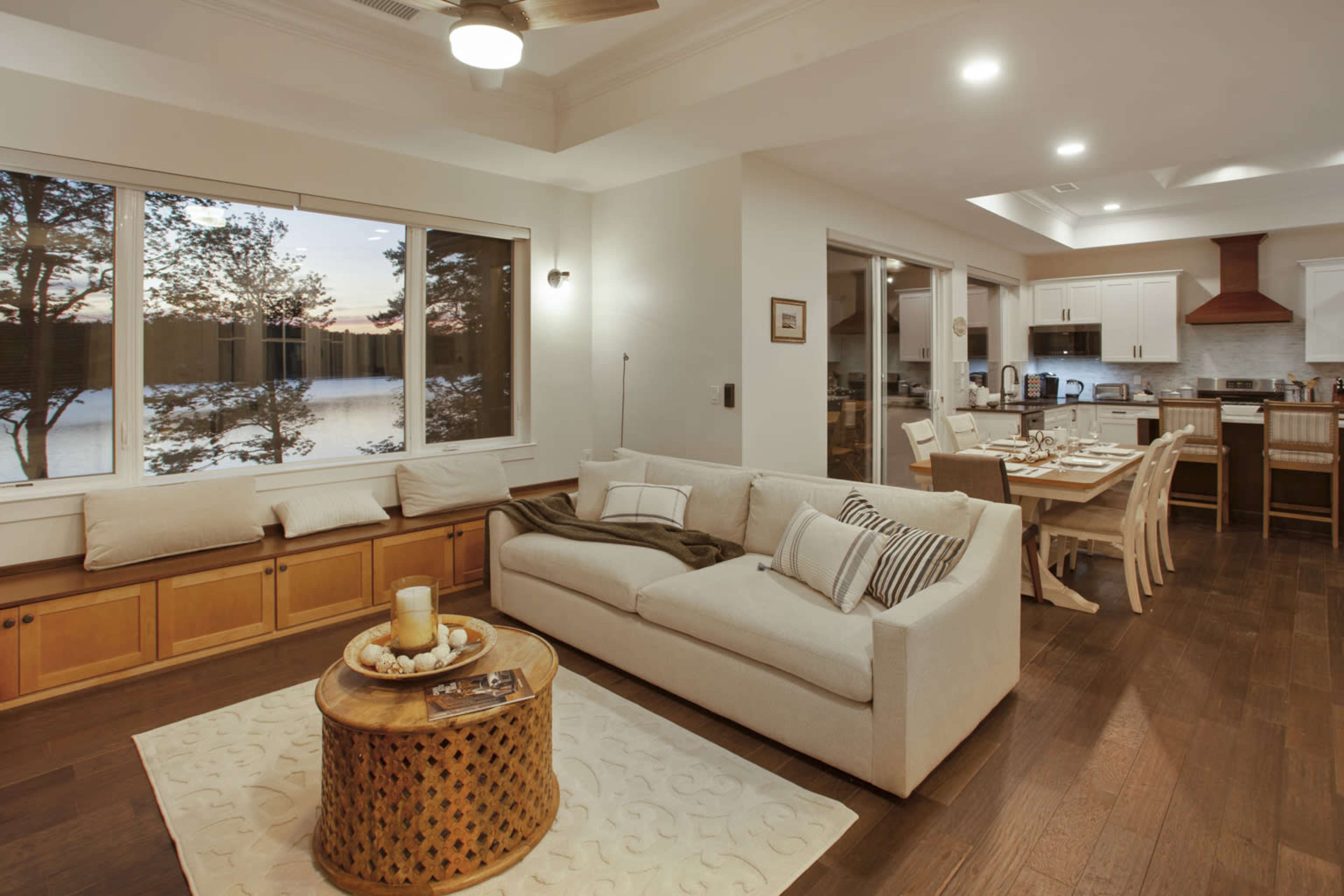 A modern living space featuring a beige sofa, a round wooden coffee table, a dining area, and large windows with a view of a lake at sunset.