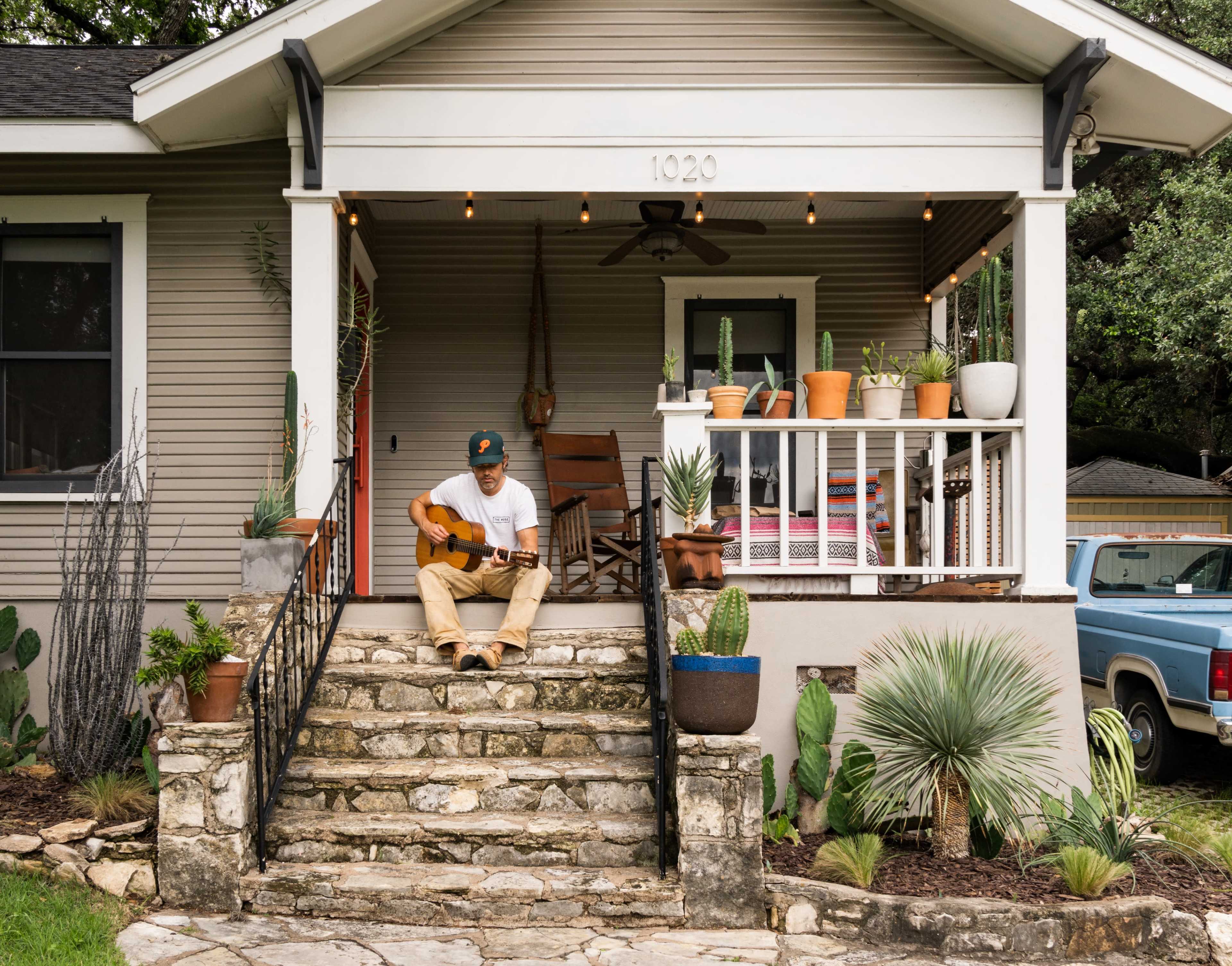A man sits on the front steps of a house, playing guitar among potted plants and decorative rocks.