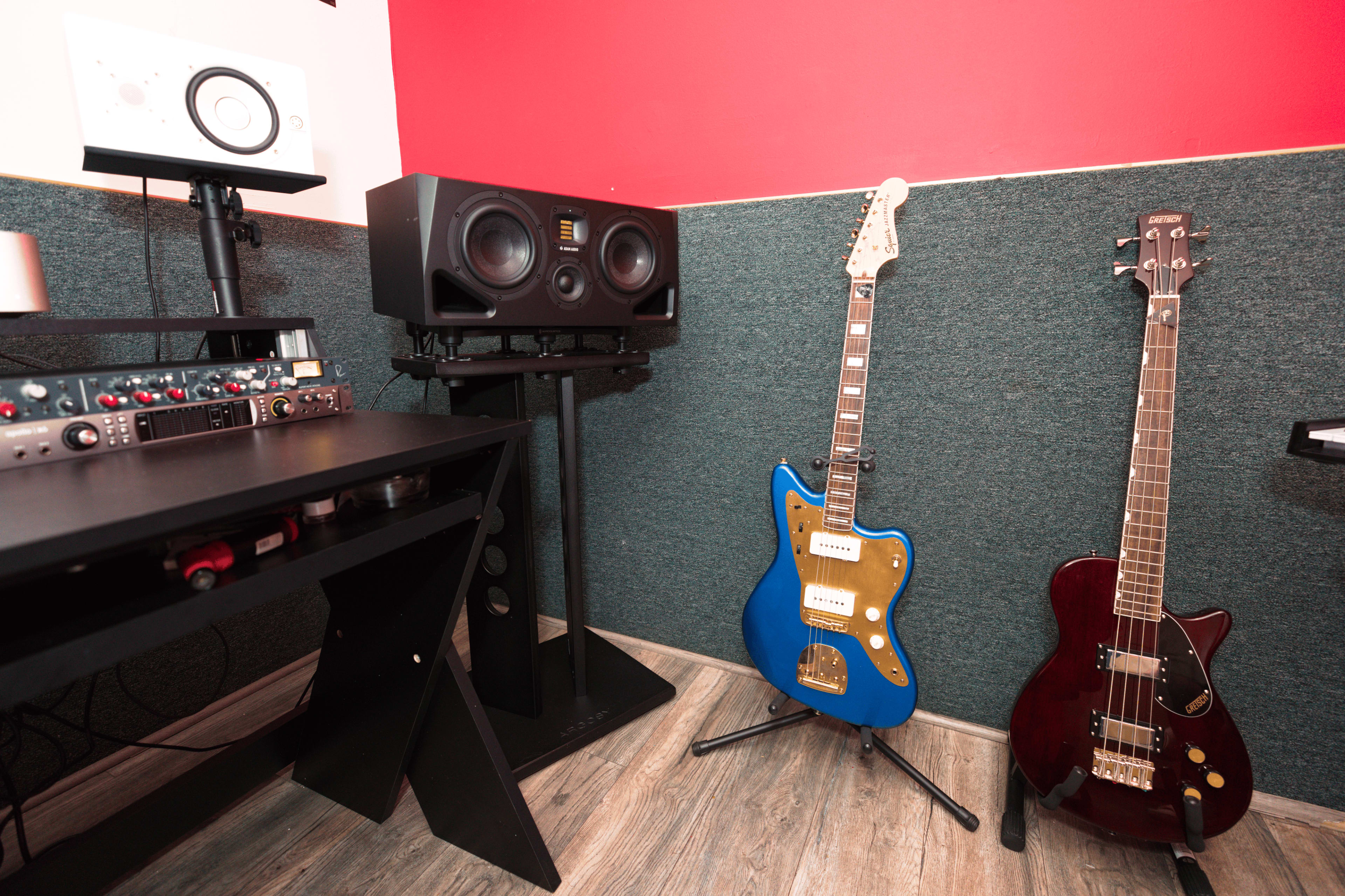 The image shows a corner of a music studio featuring two guitars, a bass guitar, a pair of studio monitors, and audio equipment on a stand.