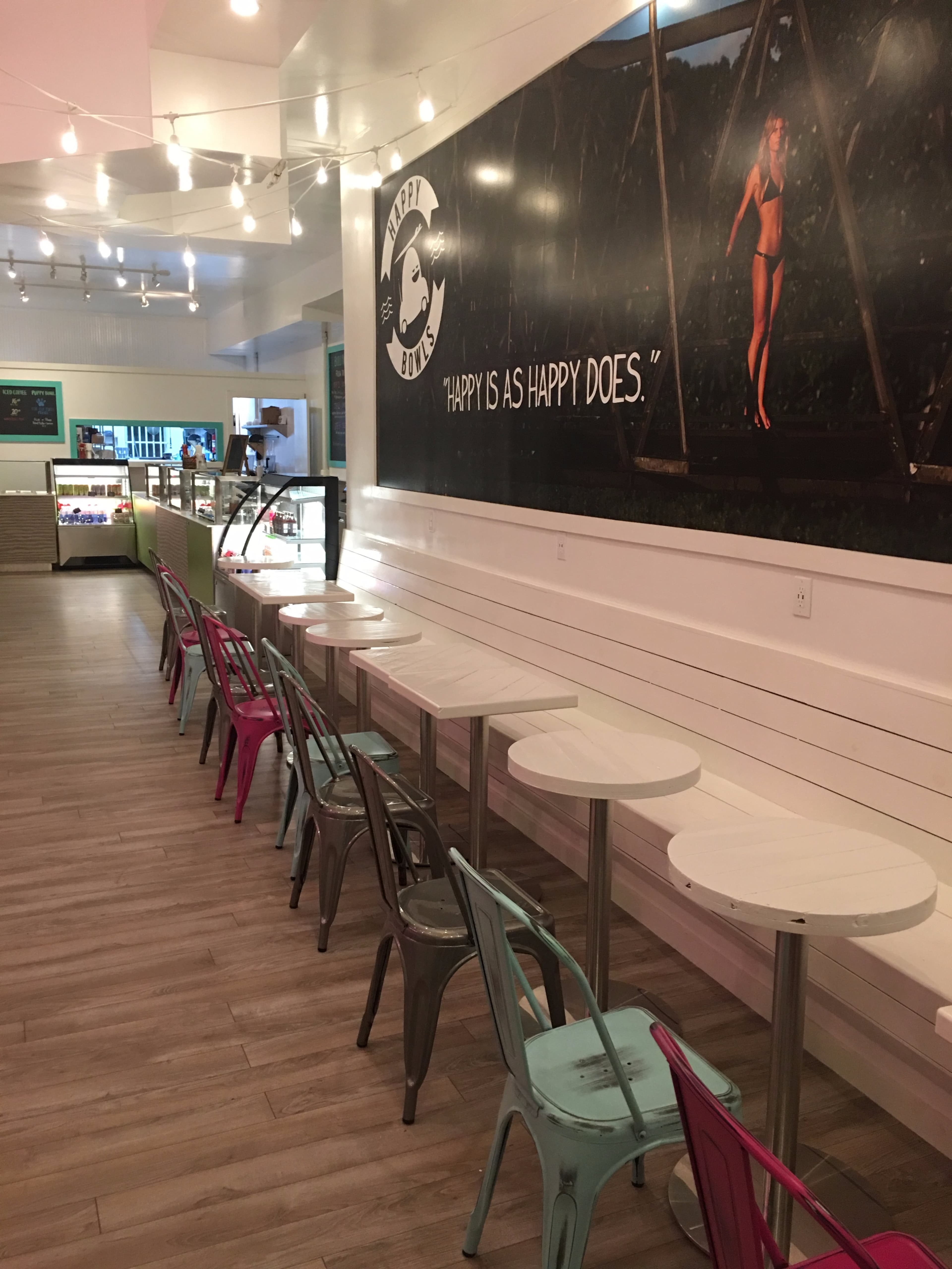 The image shows a brightly lit café interior featuring multiple small white tables with metal chairs in pink and gray, and a large wall mural with the quote "HAPPY IS AS HAPPY DOES."