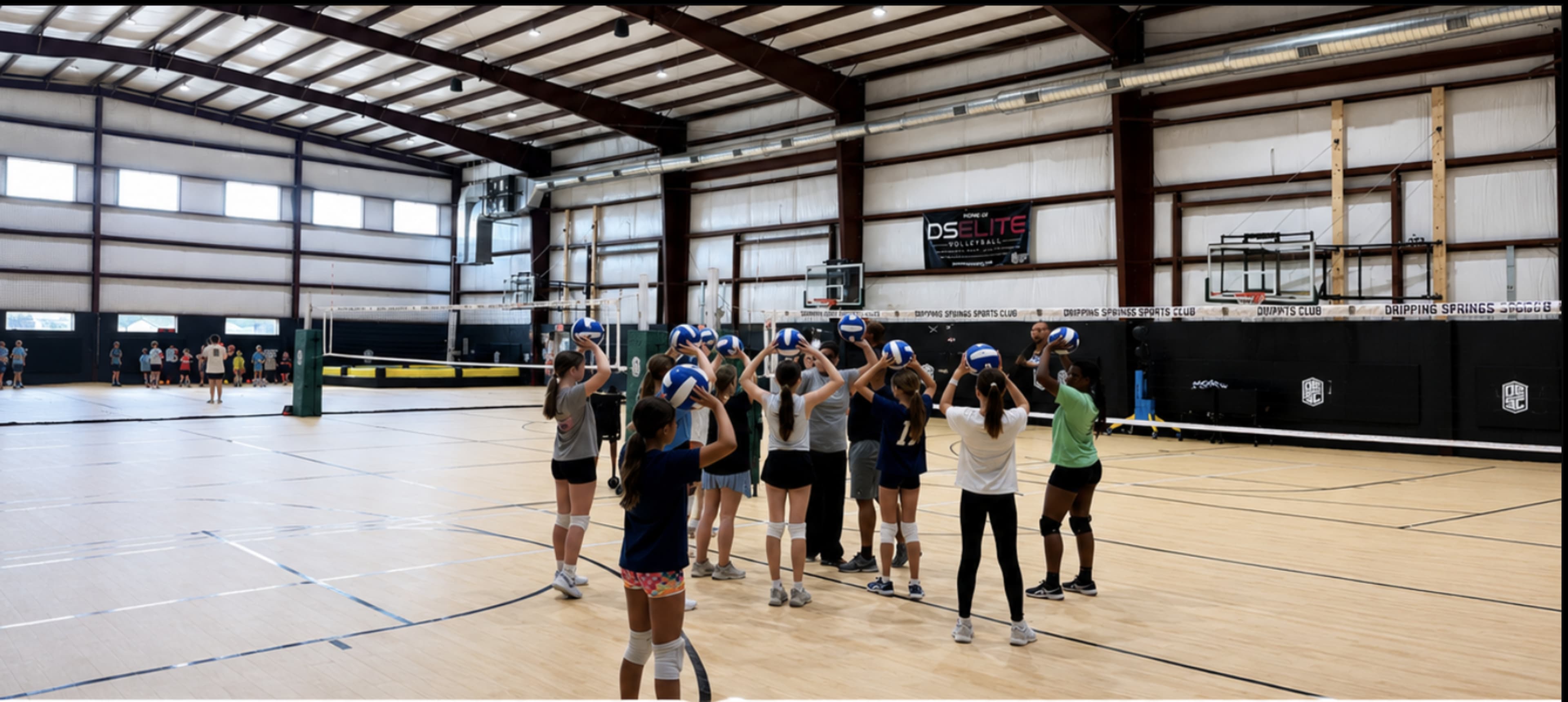 A group of young athletes is practicing volleyball drills indoors in a gym with multiple courts and basketball hoops.