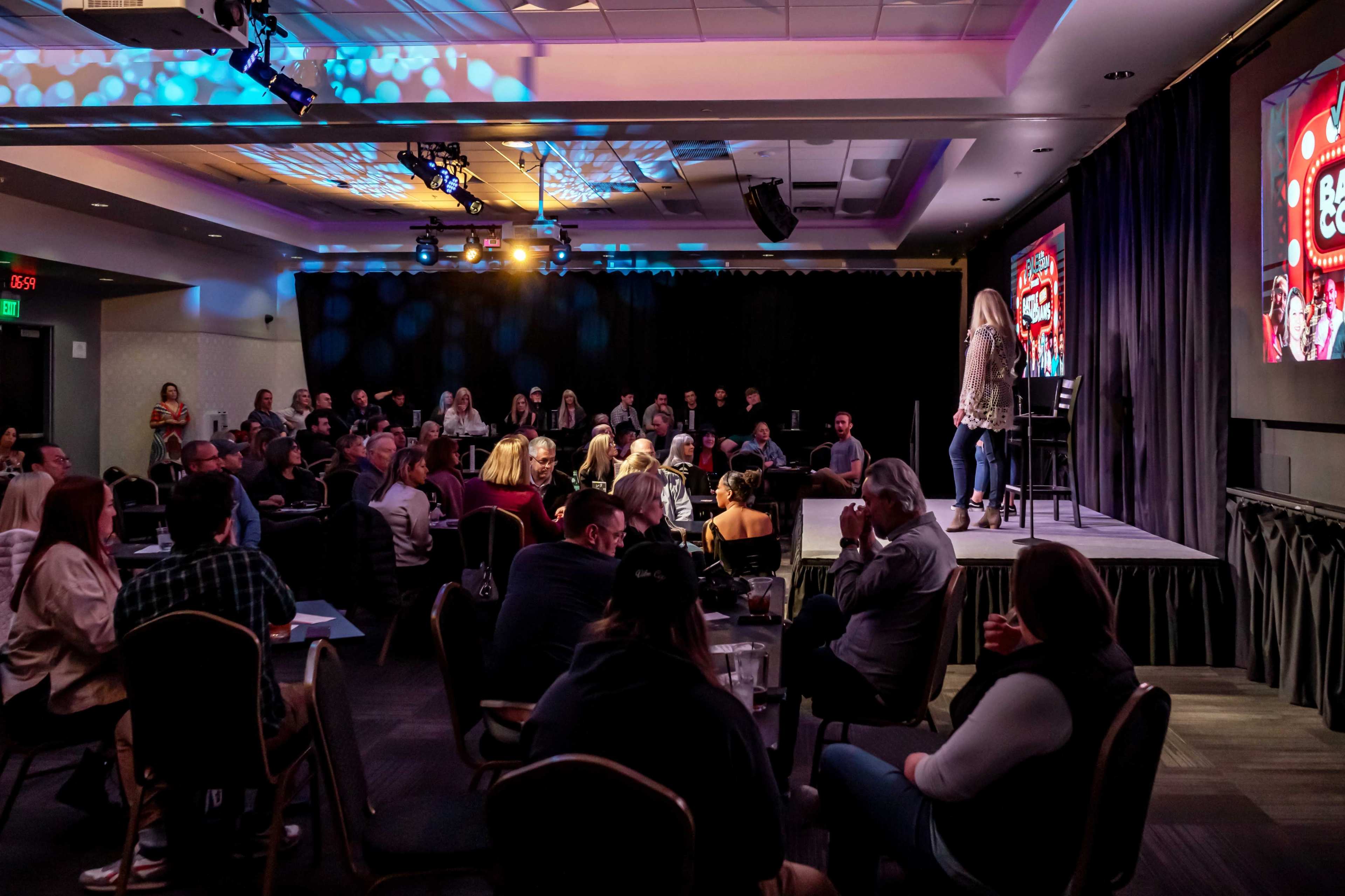 A comedian performs on stage in a well-attended comedy club, with an audience seated around tables.