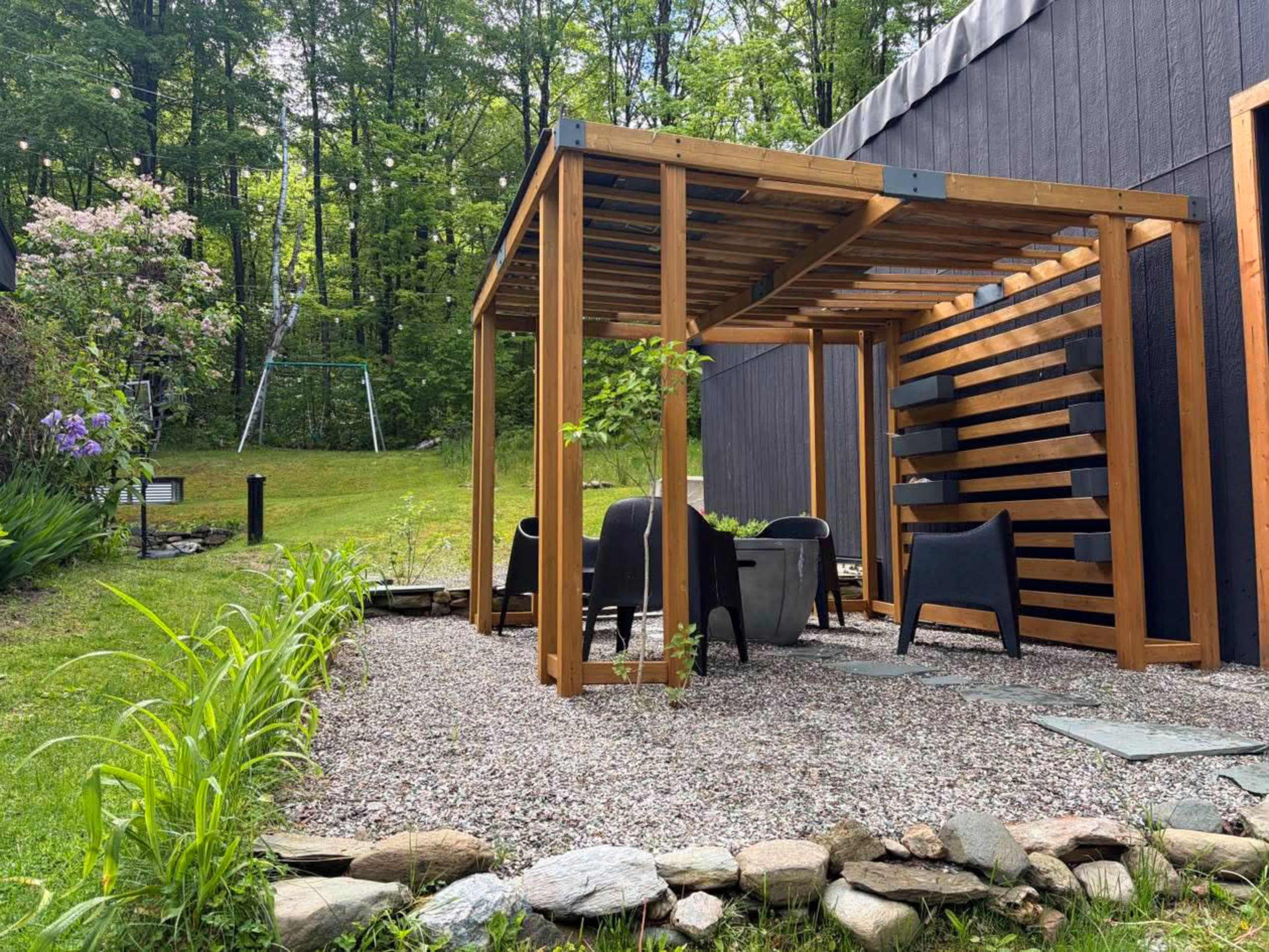 The image shows a wooden pergola structure in a garden with gravel, surrounded by greenery and nearby seating.