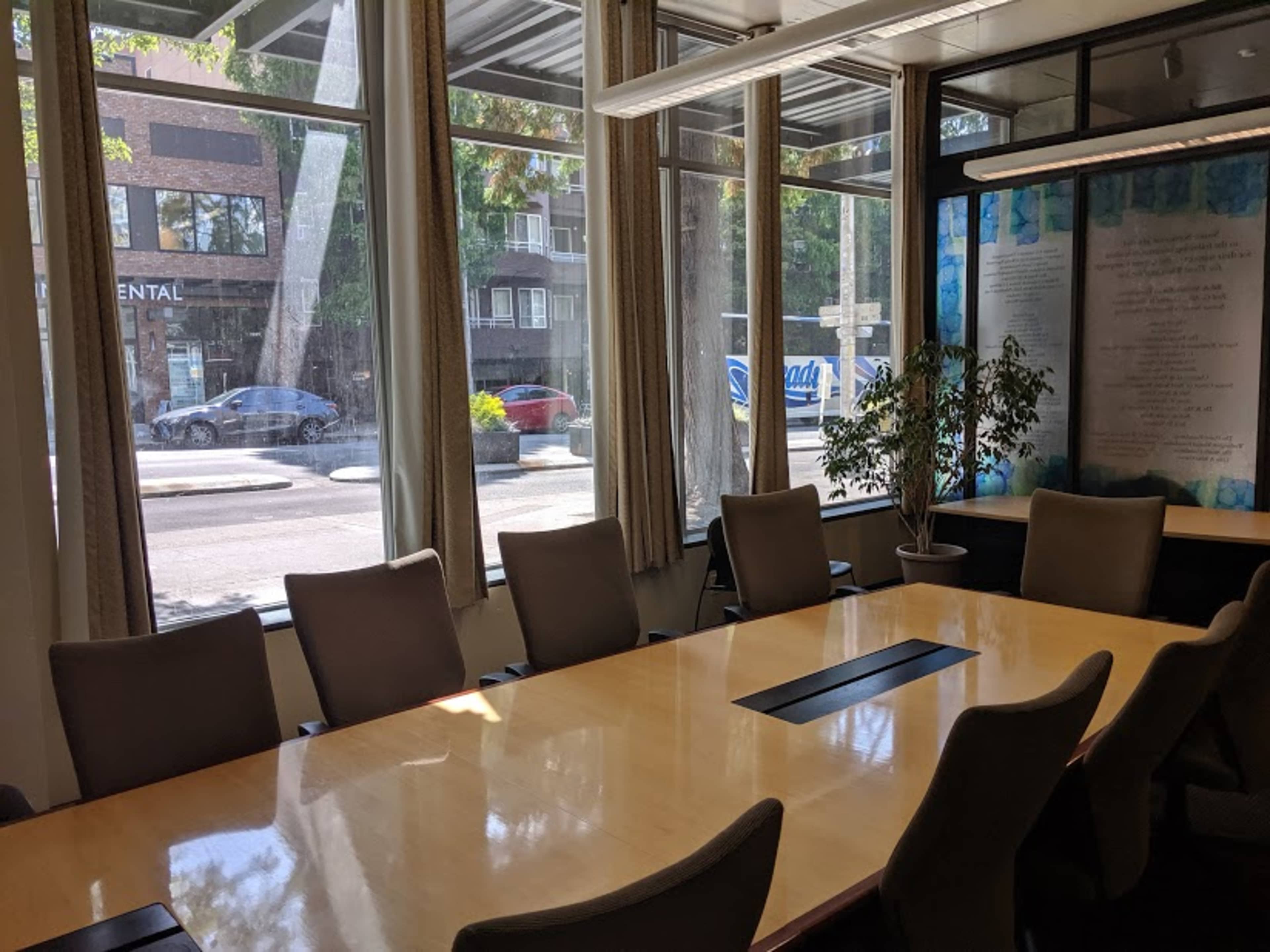 A conference room with a long wooden table surrounded by chairs, large windows letting in natural light, and a view of the street outside.