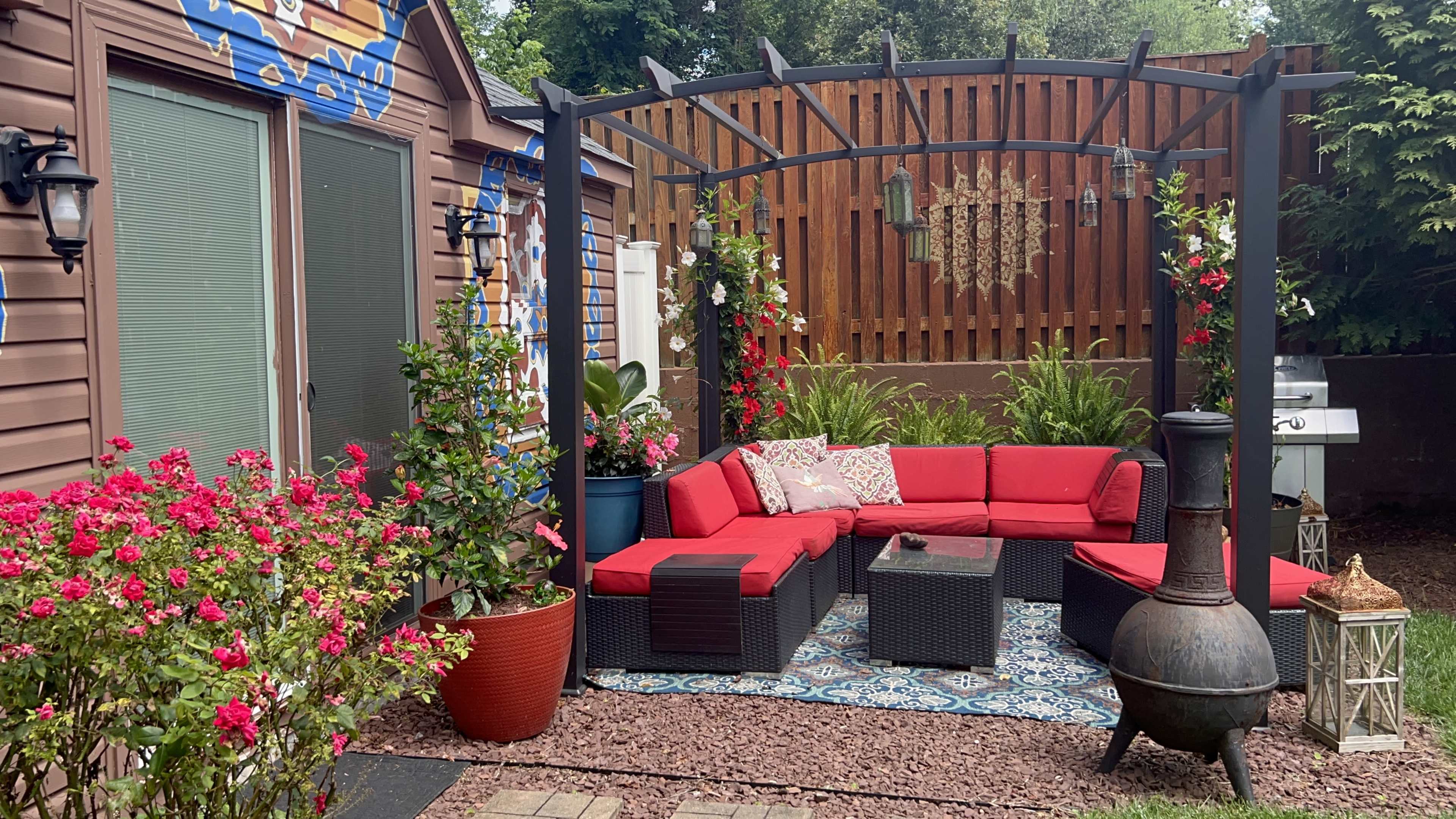 A cozy outdoor seating area with a red sectional sofa, a coffee table, decorative plants, and a barbecue grill in a landscaped yard.