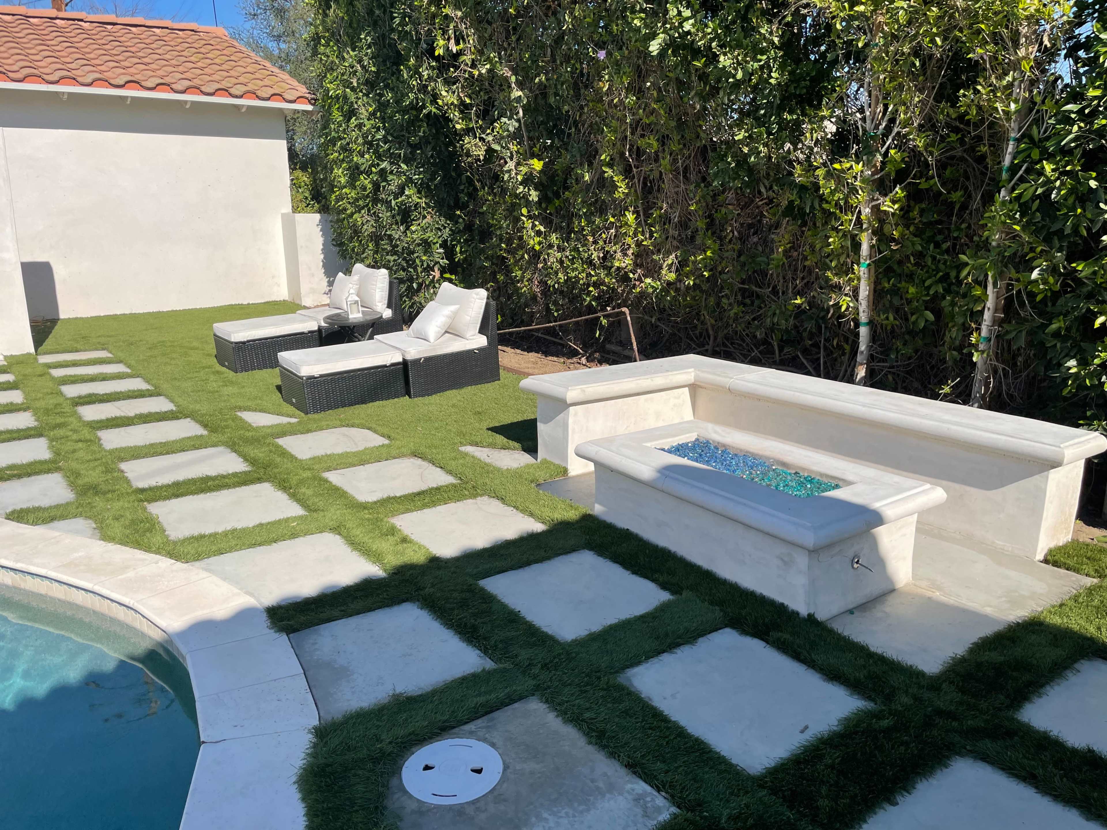 A landscaped backyard featuring a small pool, a seating area with white cushions on black wicker furniture, and a stone bench beside a rectangular water feature.