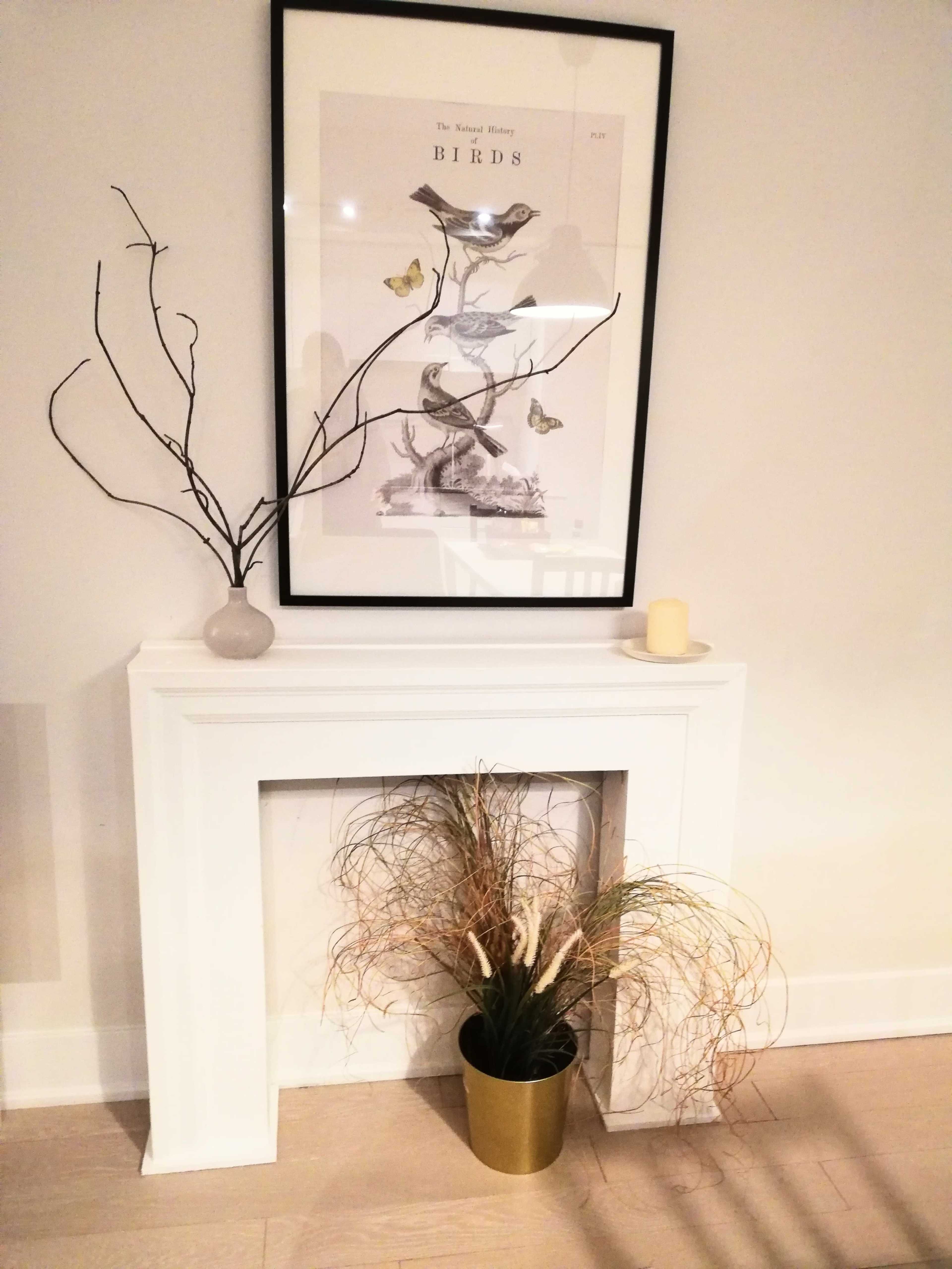 The image shows a minimalist mantel with a framed bird illustration above, a decorative vase, a candle, and a potted plant with long, wispy foliage.
