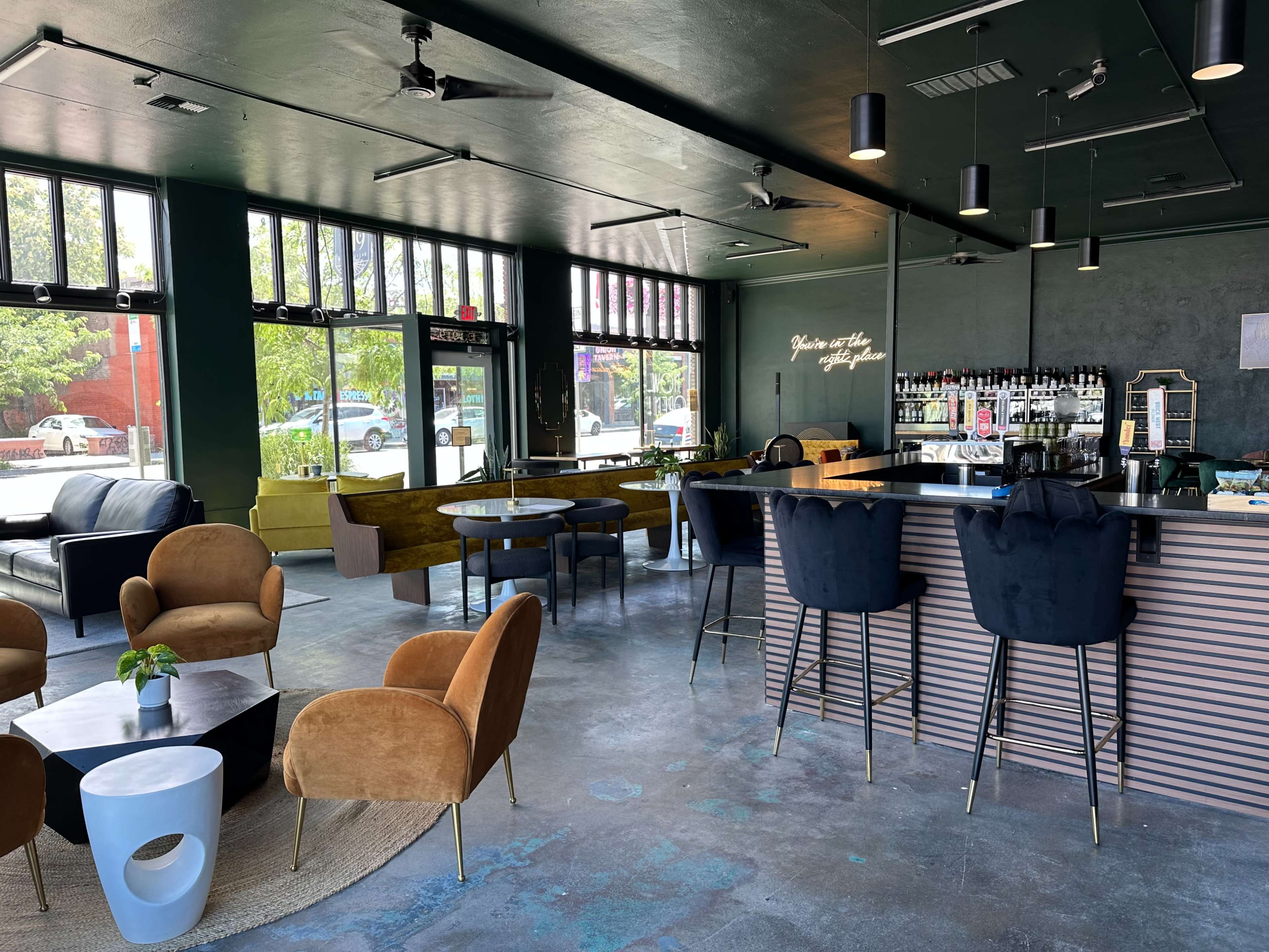 The image shows a modern café interior with a bar, seating areas, and large windows letting in natural light.