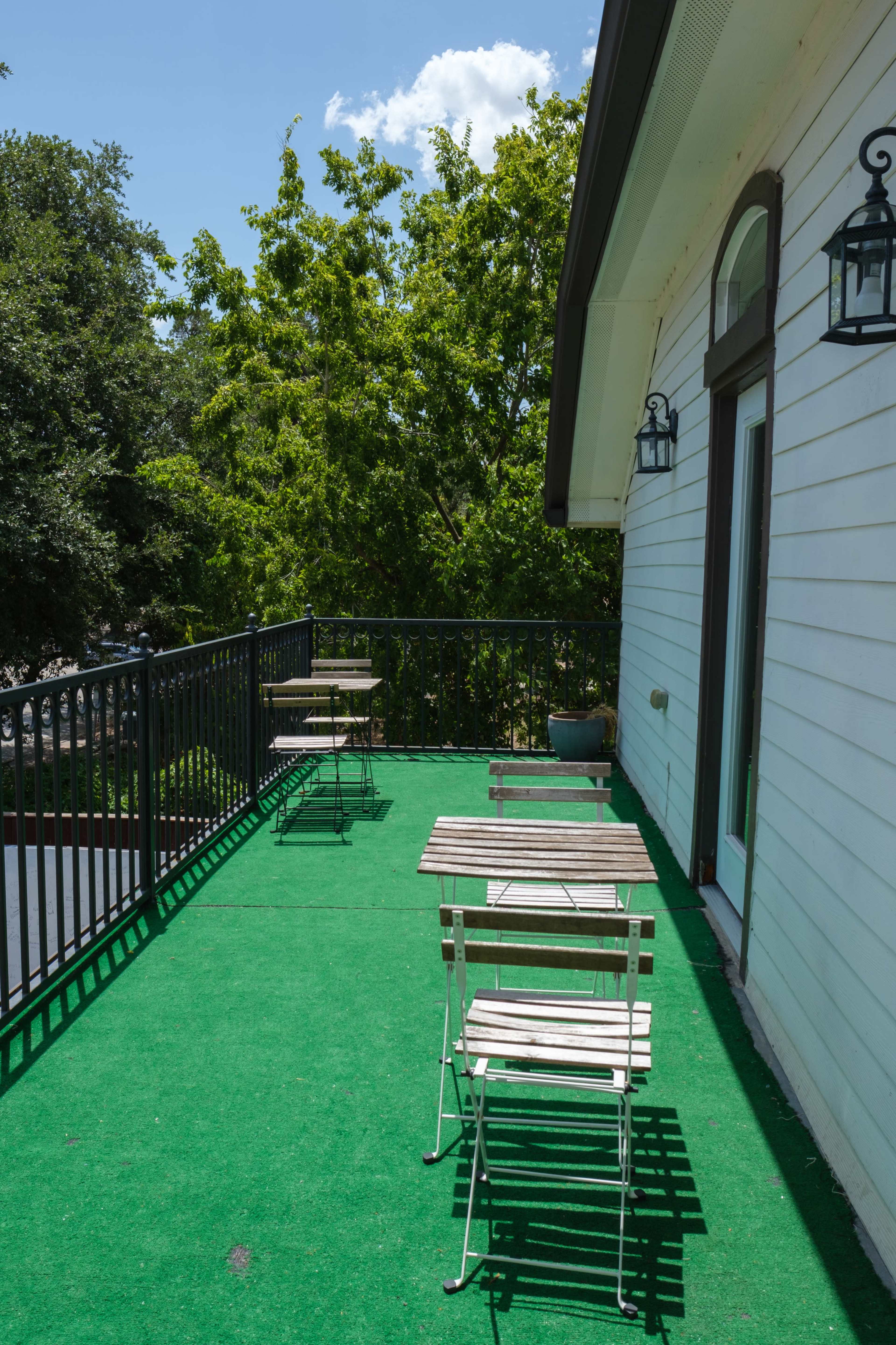 A balcony with green turf flooring and metal folding chairs set against a white wall and surrounded by trees.