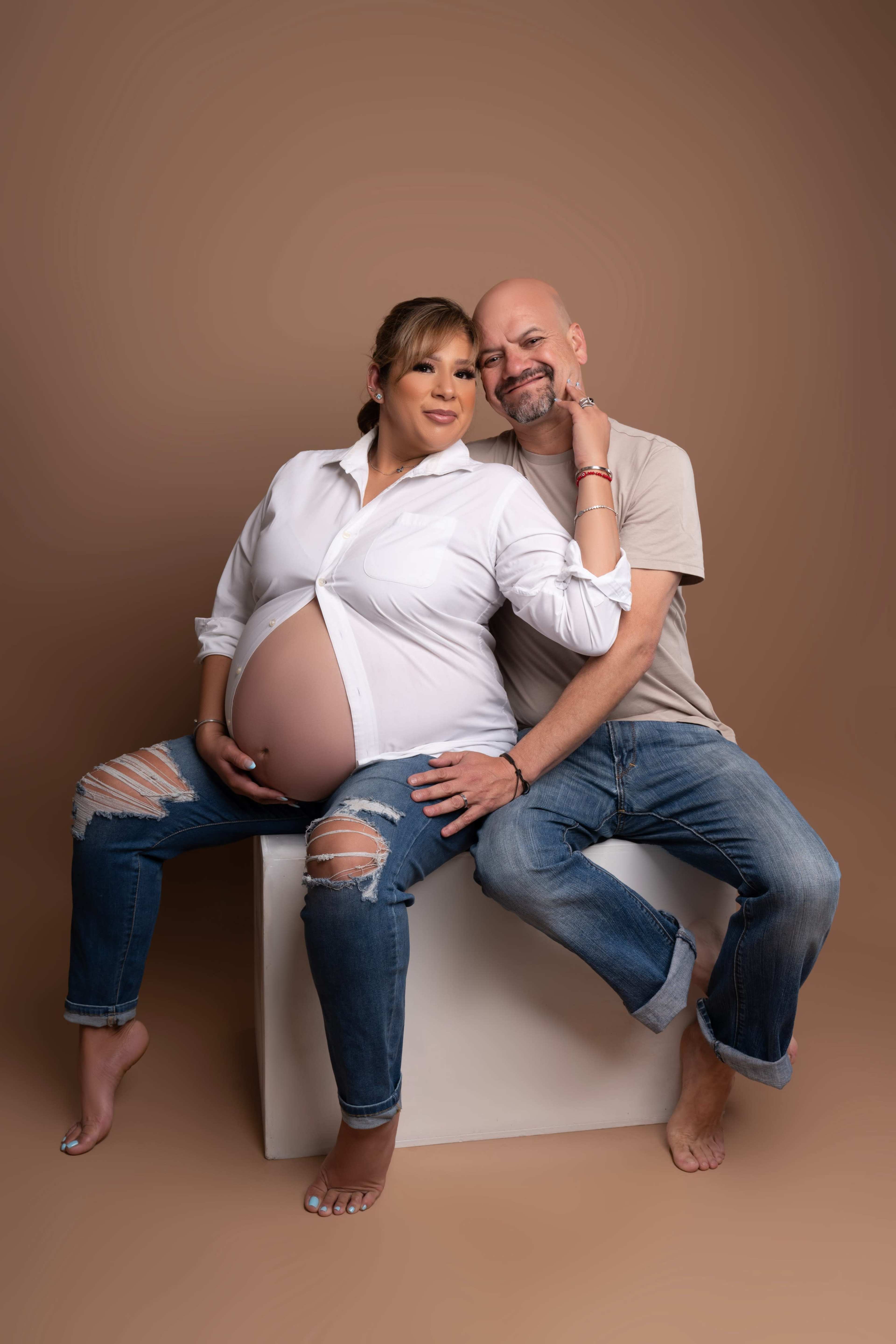A pregnant woman in a white shirt and a man in a beige shirt sit together on a stool against a neutral background.