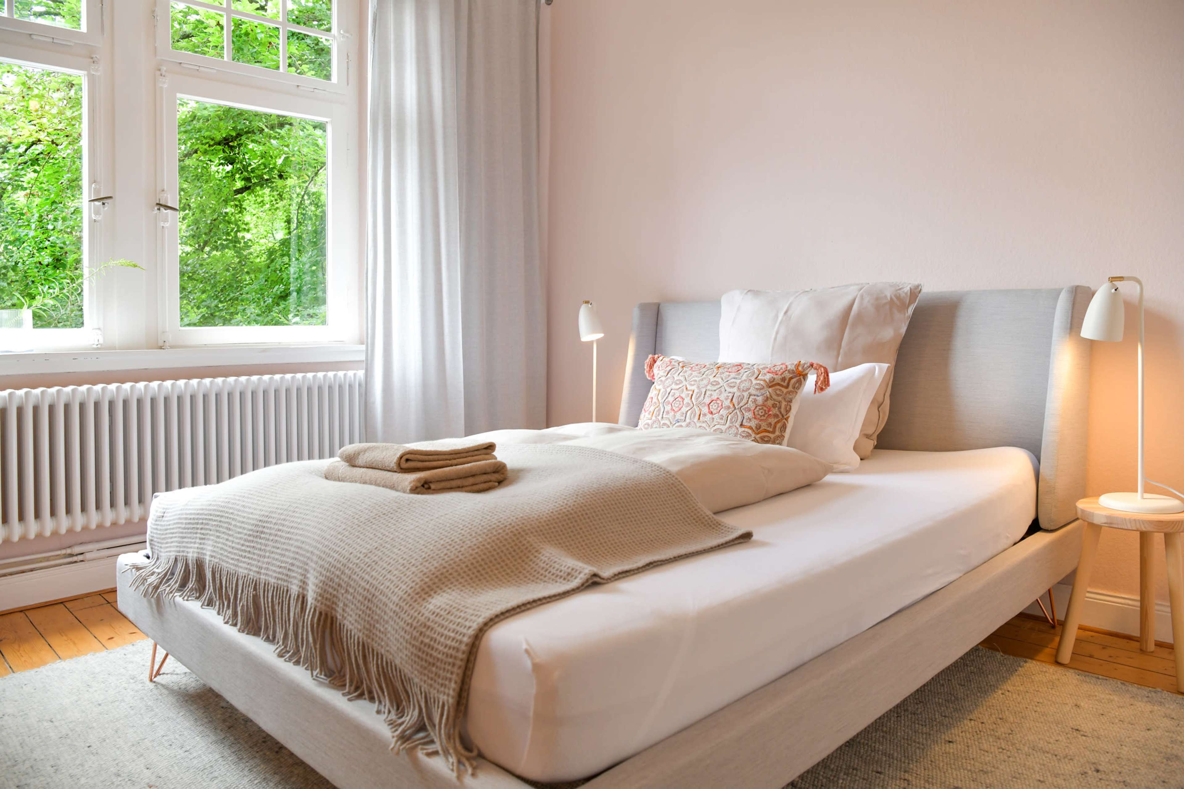A neatly made bed with a beige blanket and decorative pillow, situated in a room with large windows that overlook greenery.