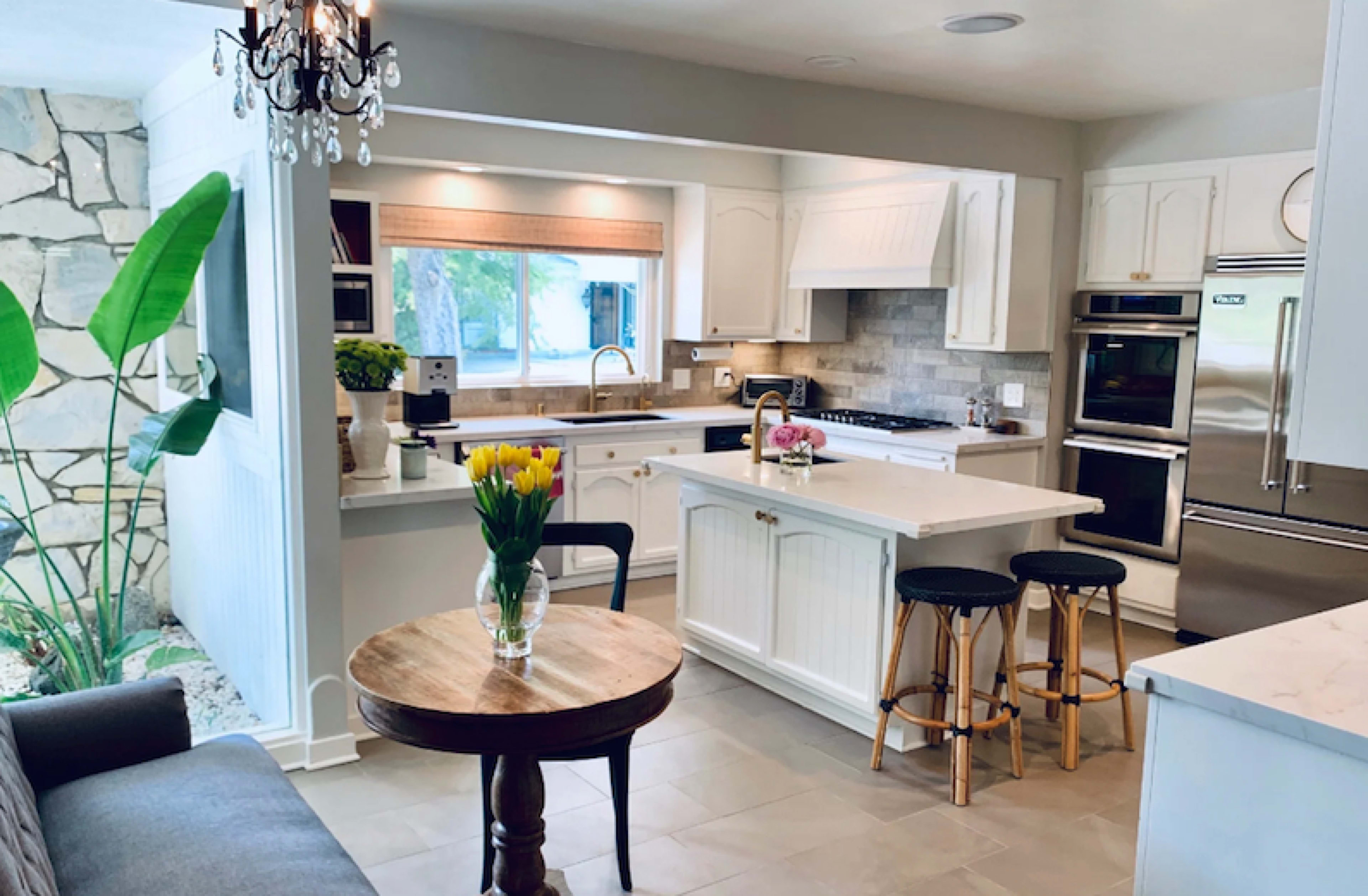 A modern kitchen features a central island with bar stools, white cabinetry, and a dining table with a vase of tulips, alongside large windows and tropical greenery visible outside.
