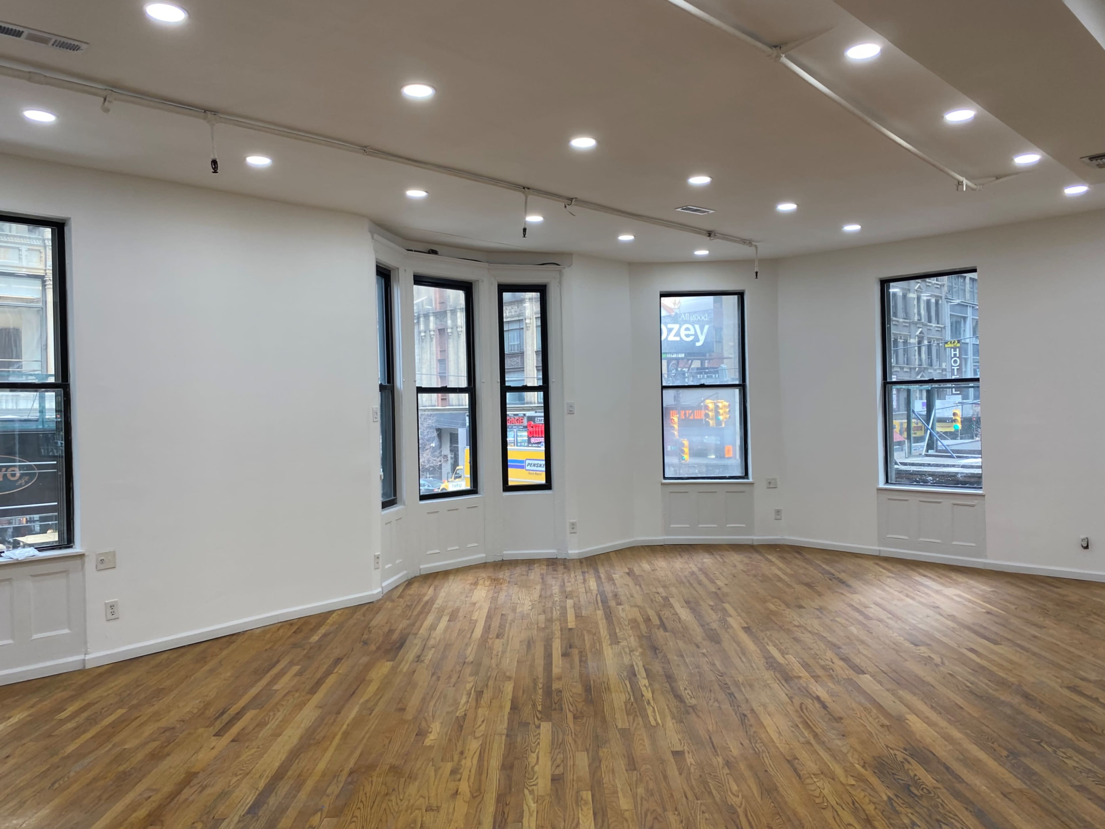 The image shows a spacious, empty room with hardwood floors and large windows that offer a view of a city street.