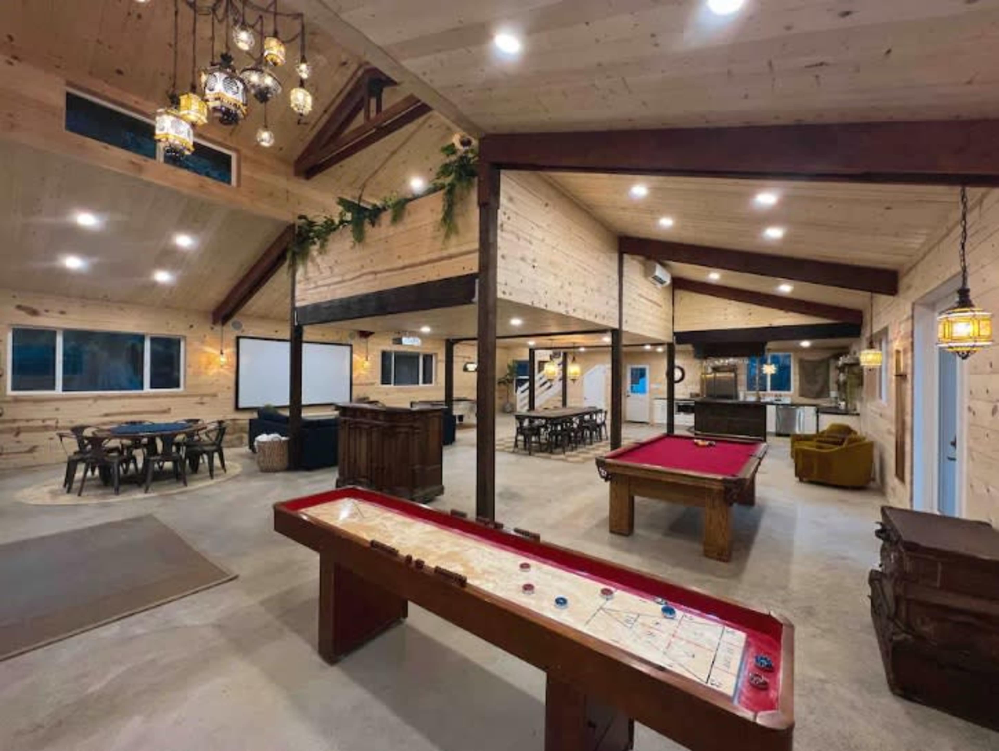 The image shows a spacious, wooden interior game room featuring a billiards table, an air hockey table, and multiple seating areas with large windows.