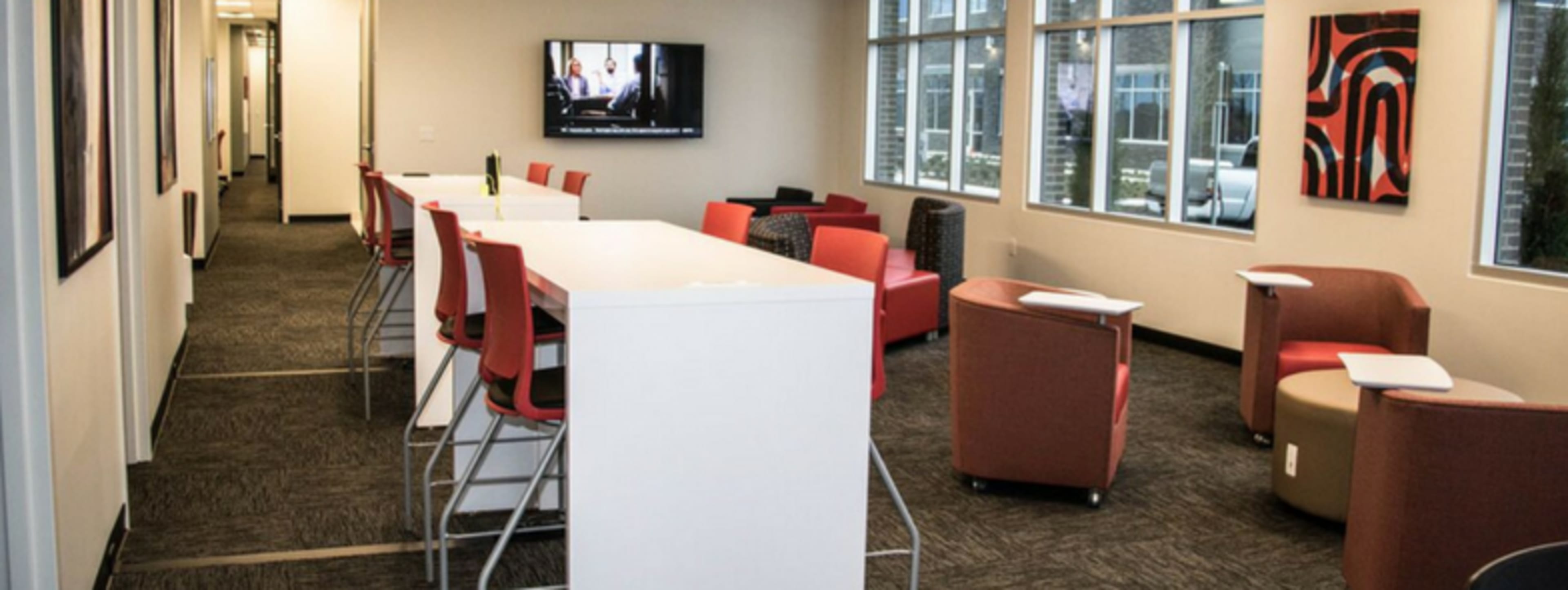 A modern shared workspace with a long white table, red chairs, and various seating arrangements along a well-lit corridor.