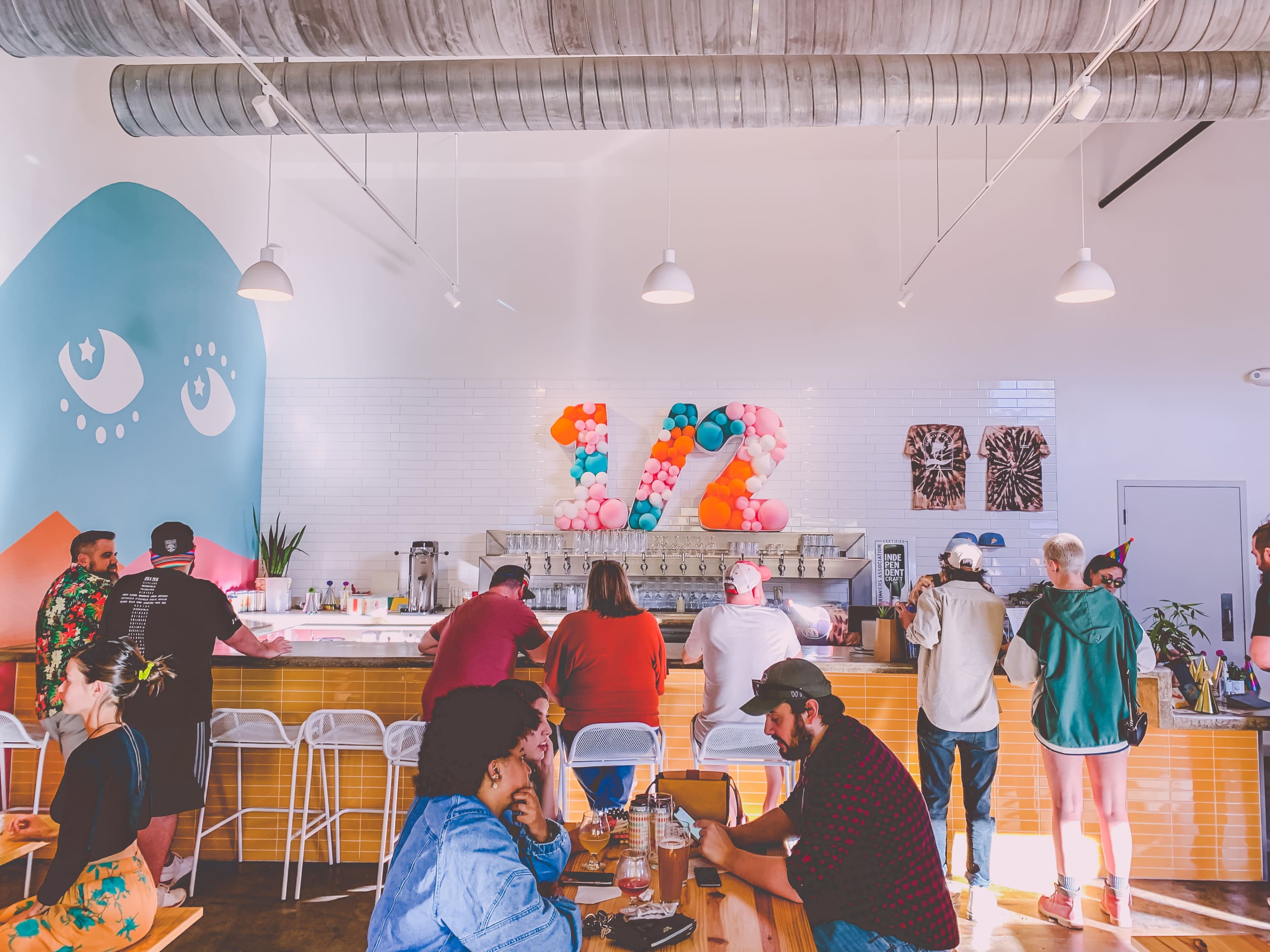 A vibrant café interior features a decorative wall with colorful balloons shaped as "12," while patrons line the counter and sit at tables.