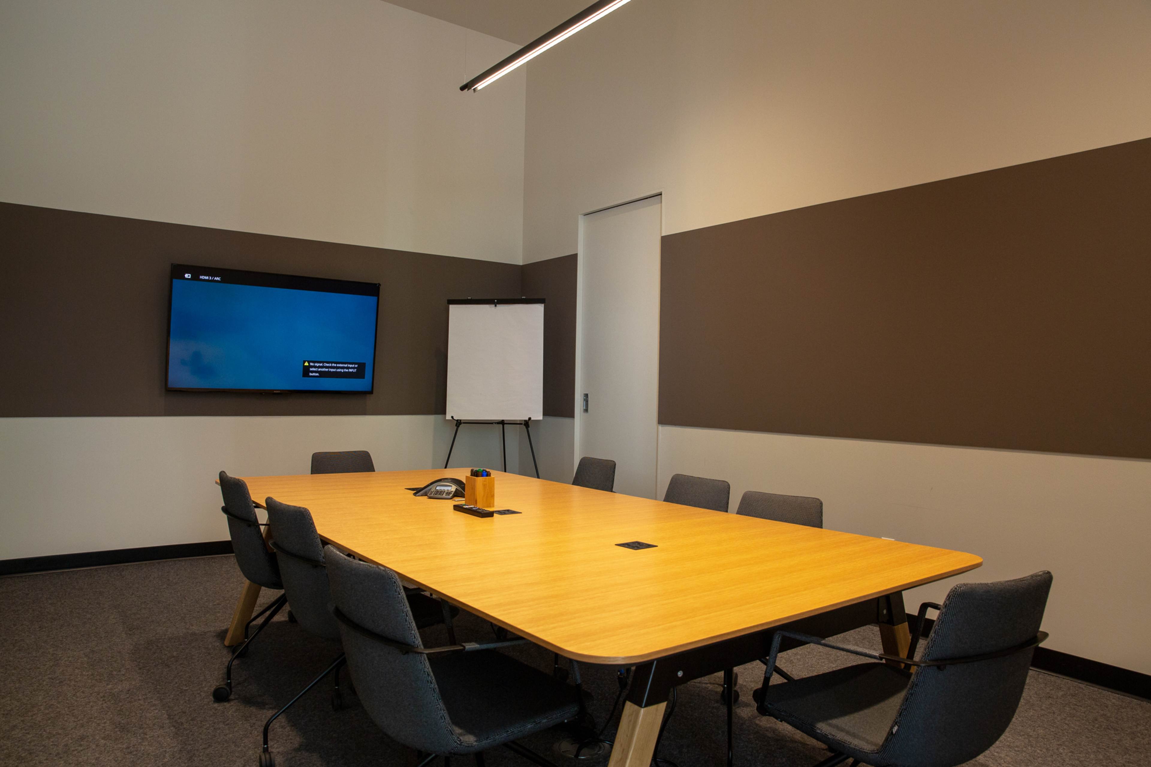 A conference room features a large wooden table surrounded by chairs, with a screen mounted on the wall and a flip chart nearby.