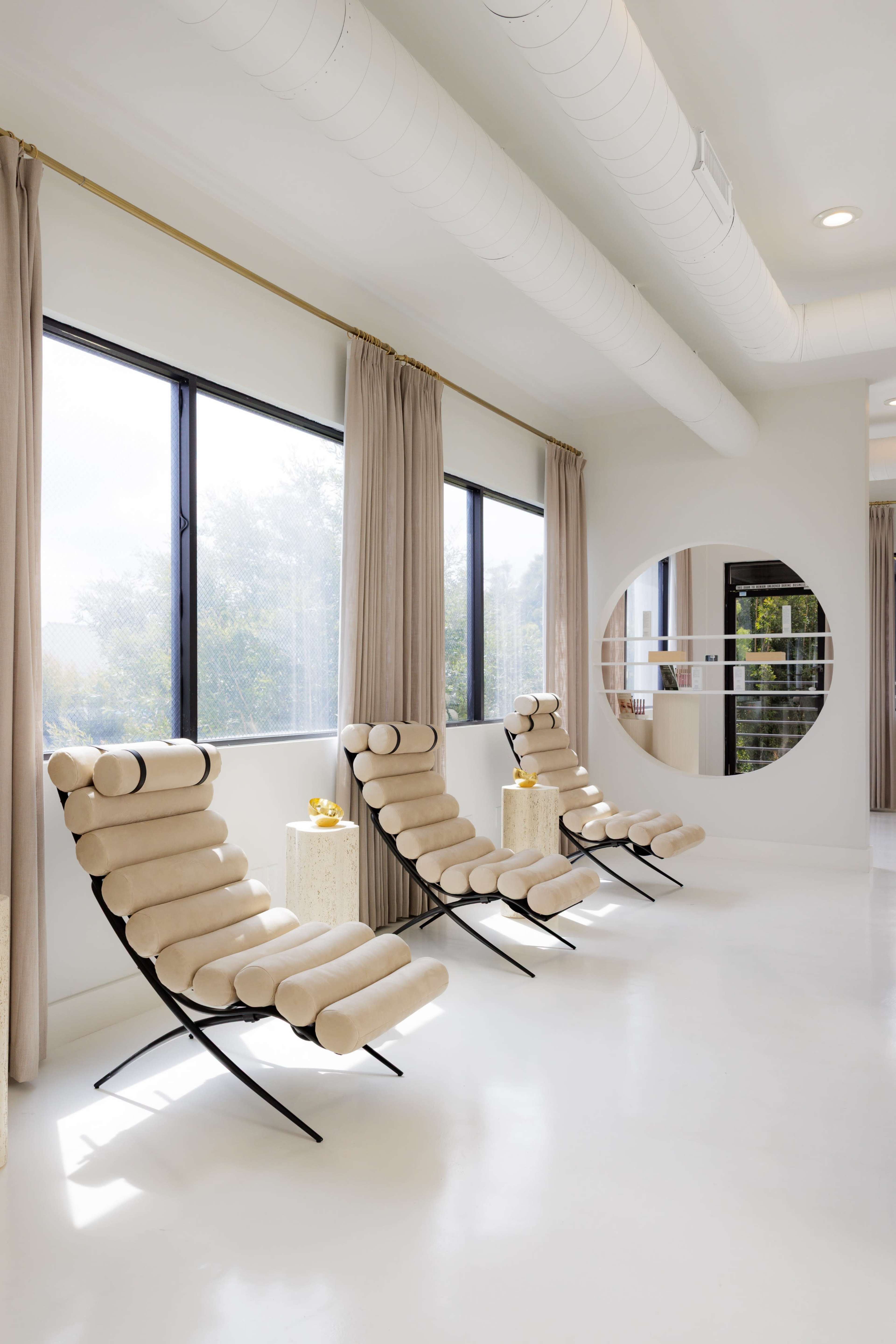 The image shows a modern interior with three beige lounge chairs facing large windows, accompanied by round mirrors and light-colored curtains.