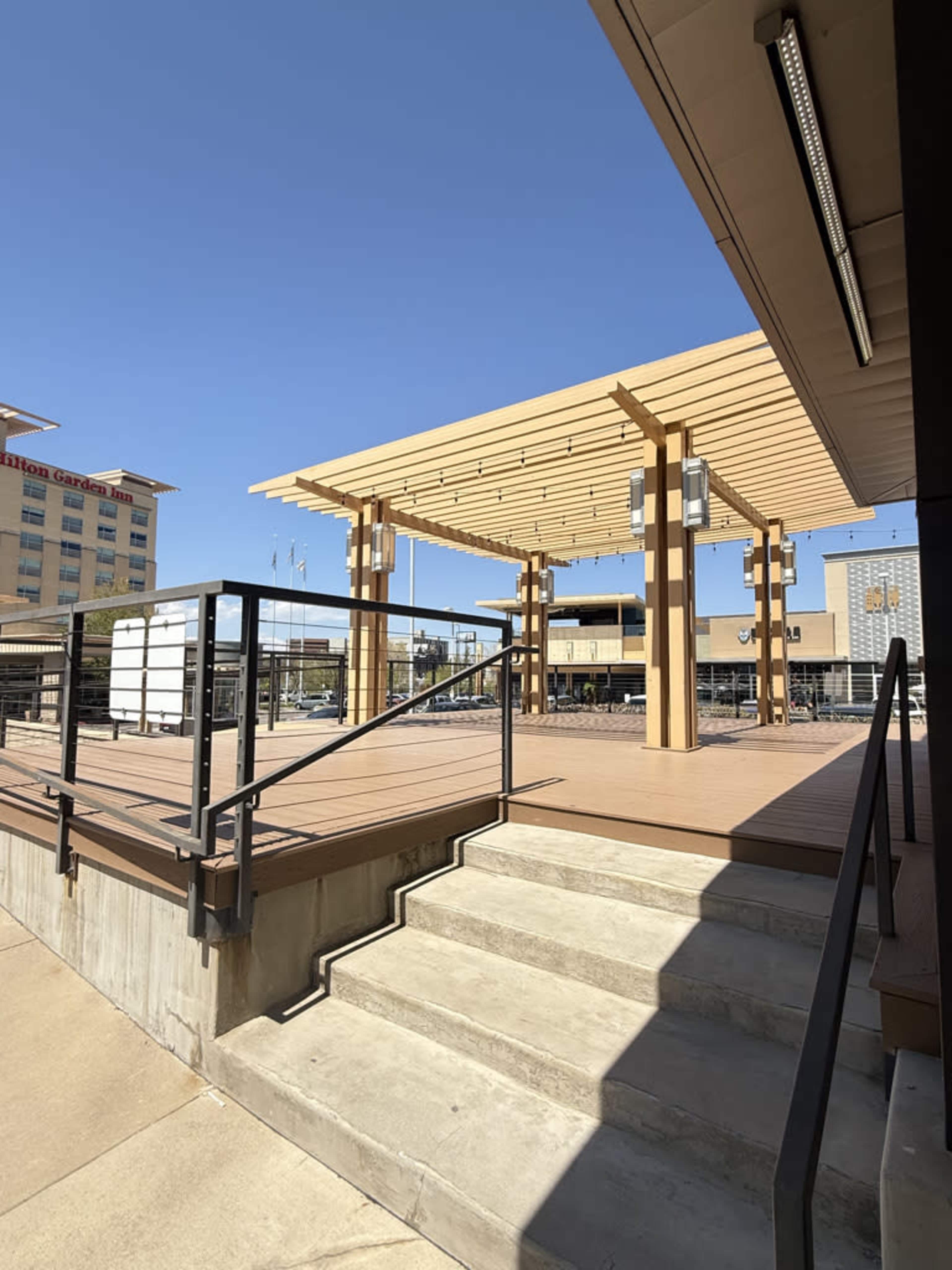The image shows a wooden deck area with a pergola structure, accompanied by stairs and a railing, under a clear blue sky.