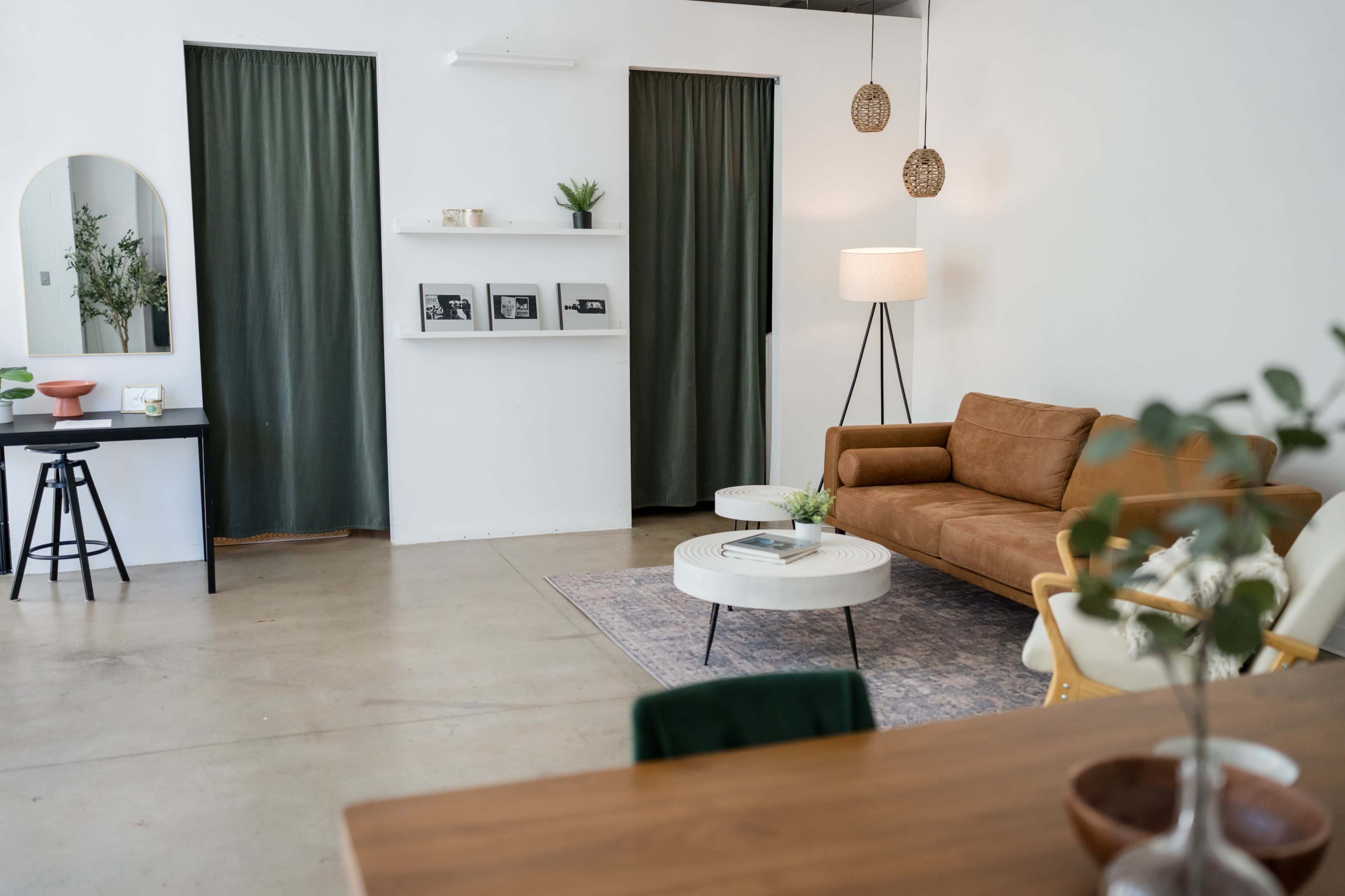The image shows a modern living room with a brown sofa, two white coffee tables, a black table with framed photos, and green curtains.