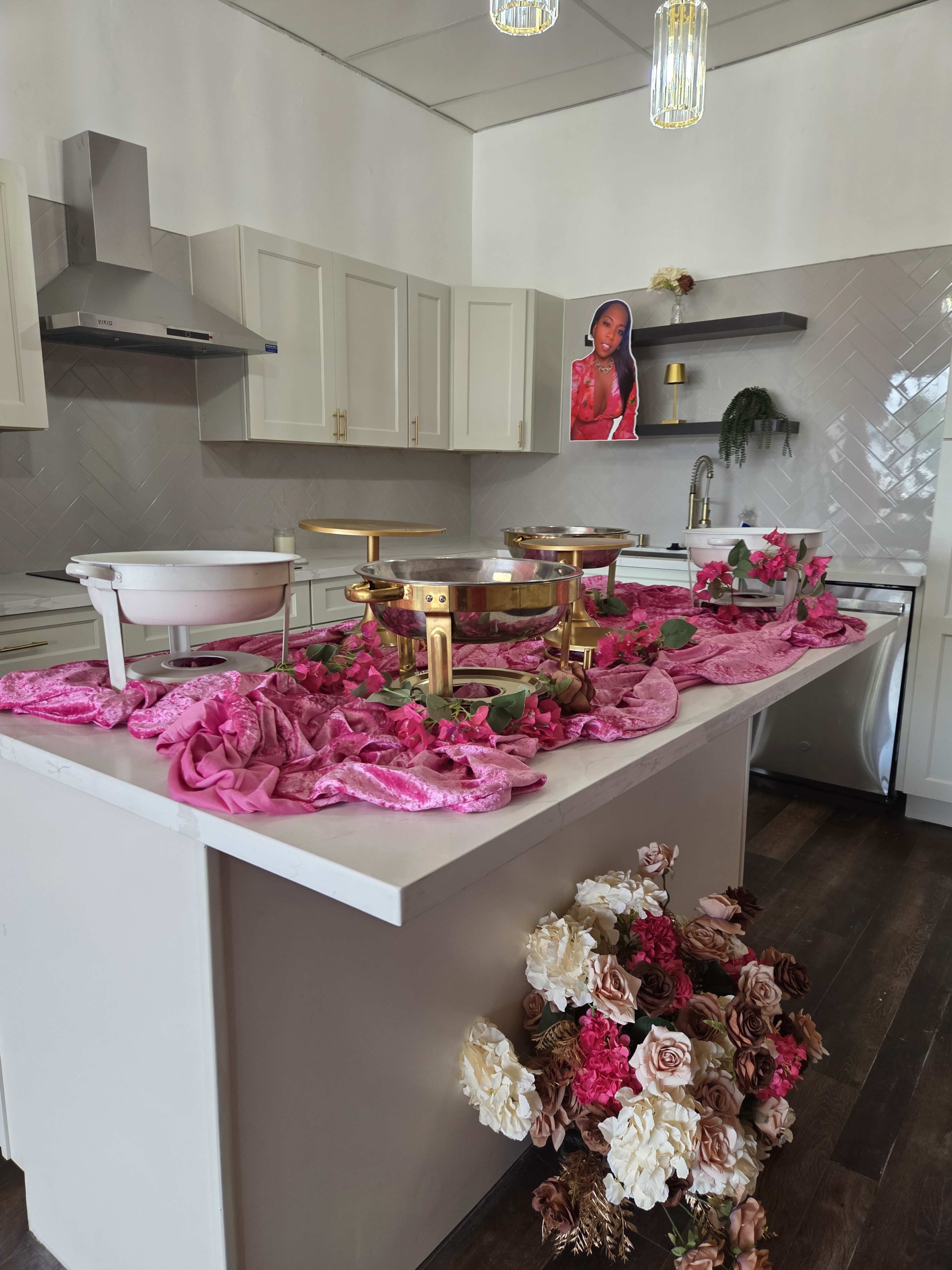 A modern kitchen features a marble island adorned with pink fabric and floral arrangements, complemented by elegant decor and a portrait on the wall.