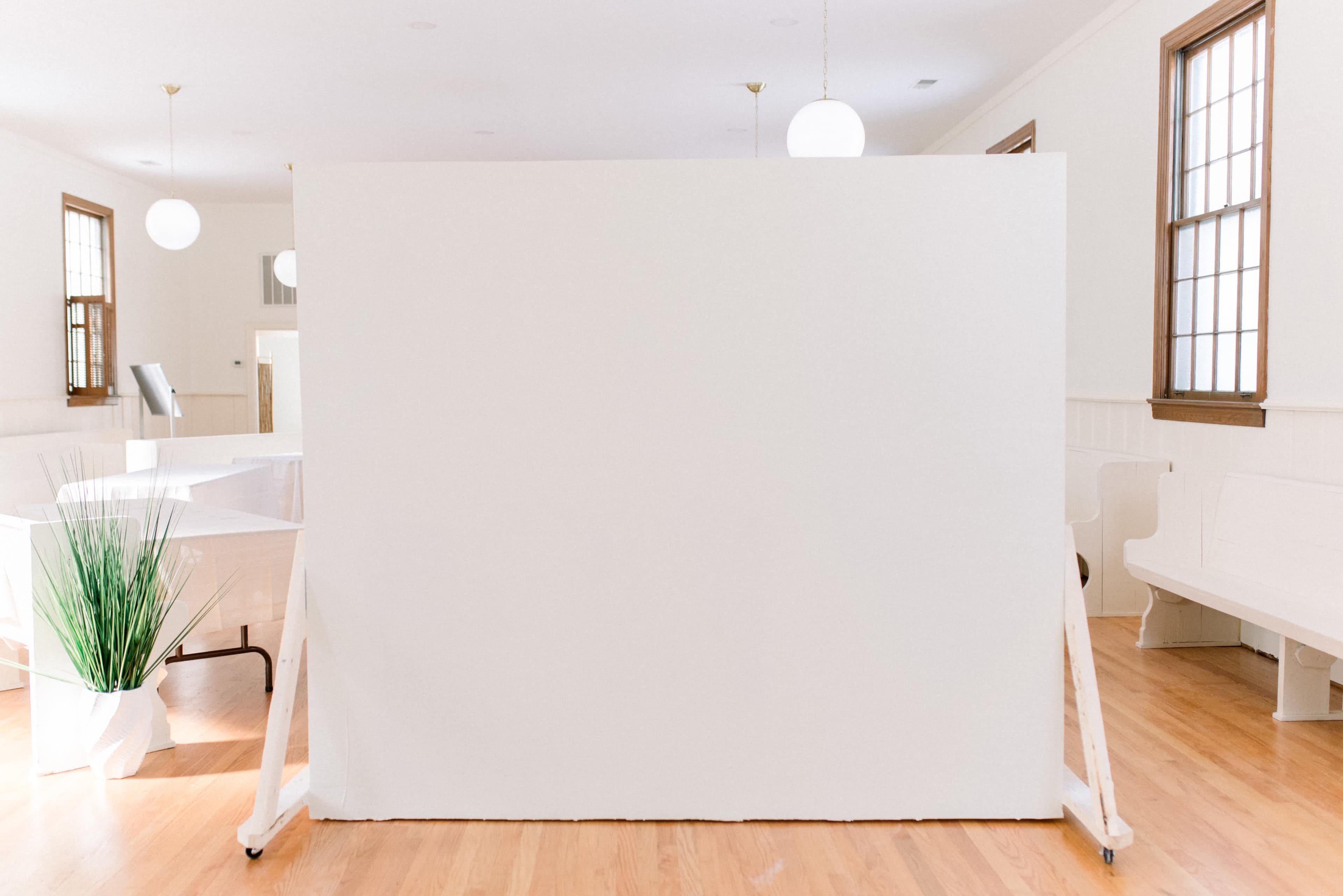 A large white board stands on a wooden floor in a bright room with windows and pendant lighting.
