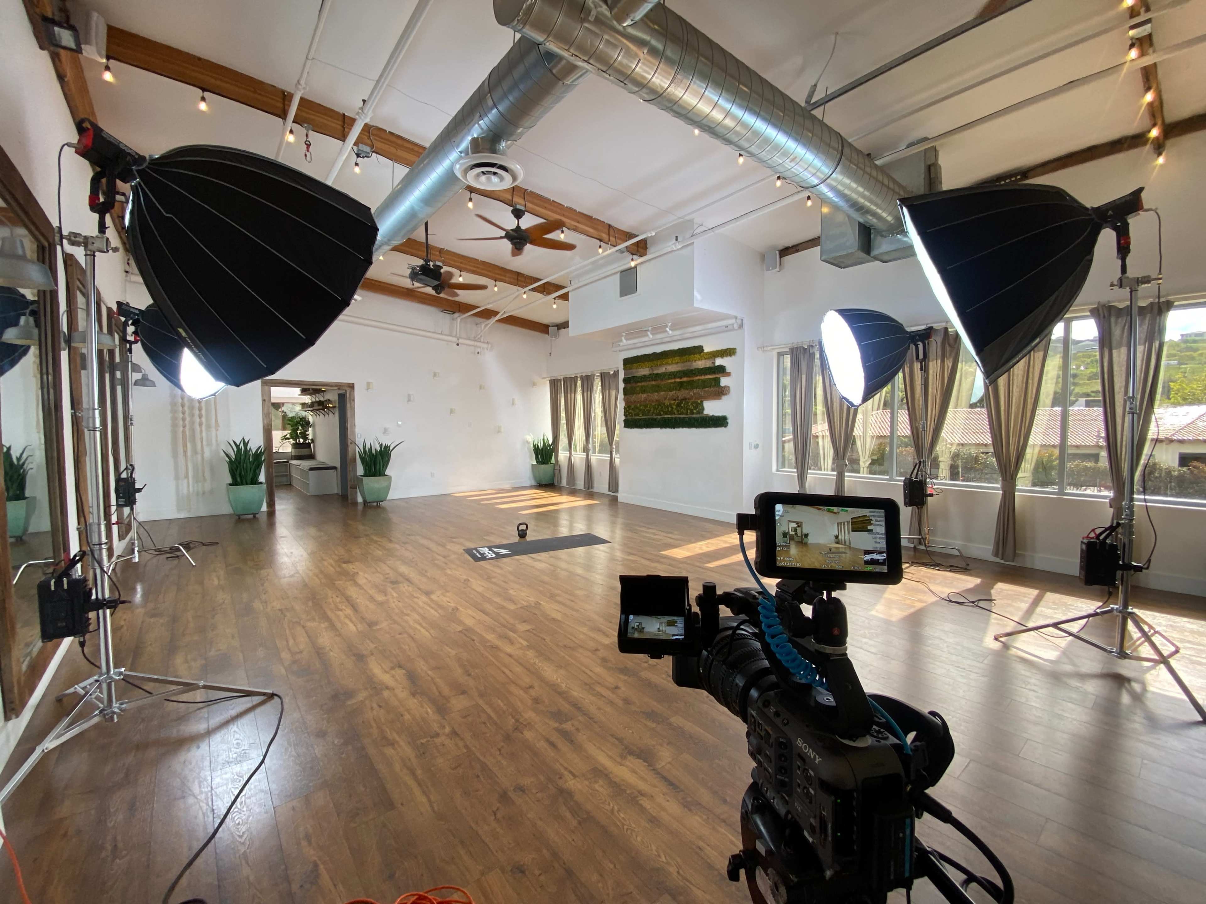 A spacious photography studio features two large softbox lights, a video camera on a tripod, and a central black mat on a wooden floor.
