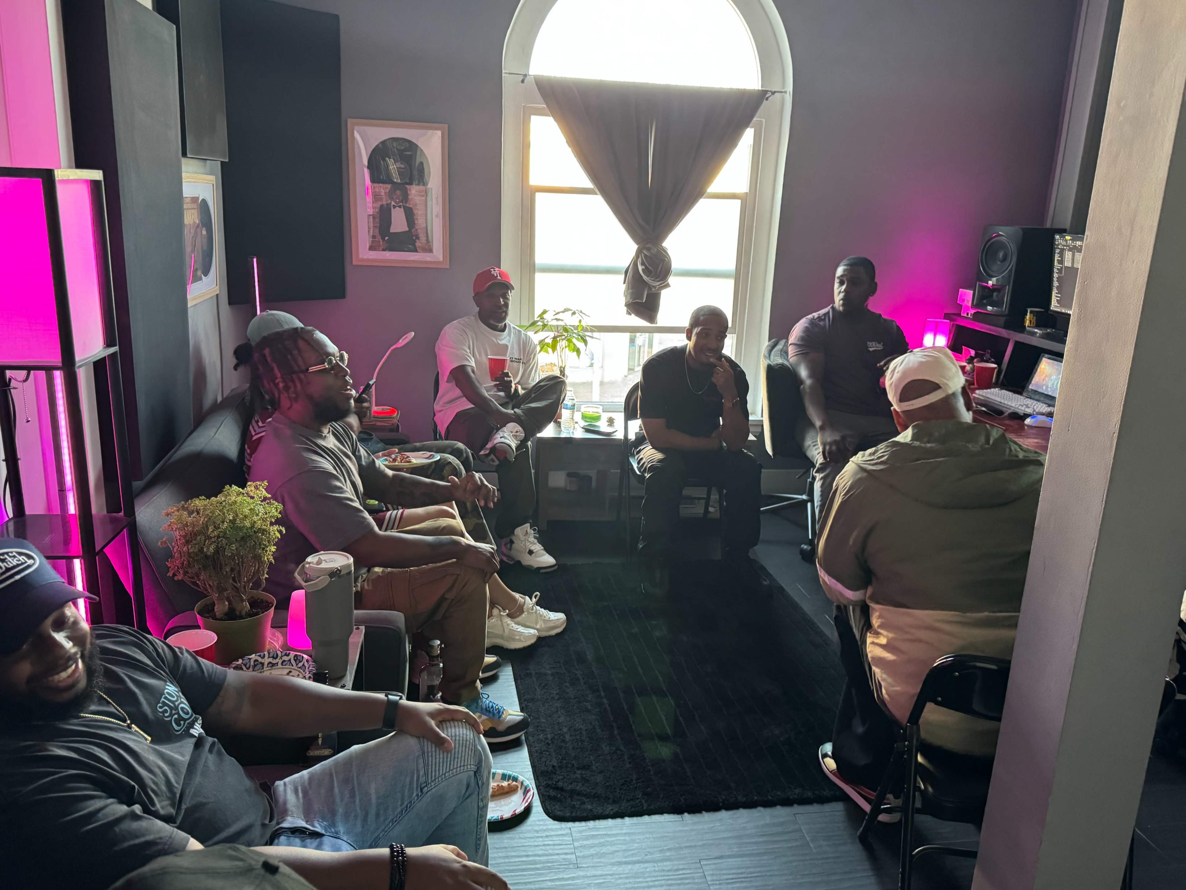 A group of seven people is seated in a well-lit room with musical equipment and a large window.