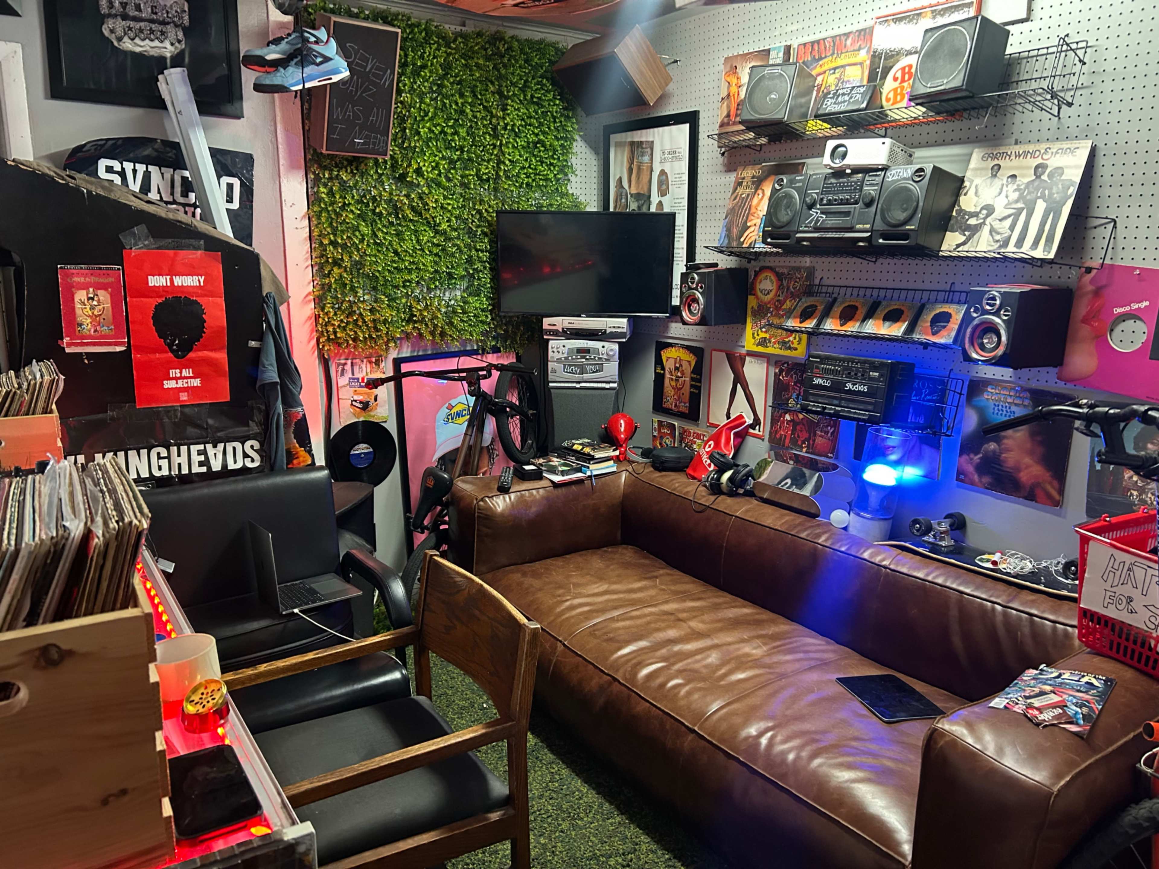 The image depicts a cozy, eclectic room filled with vintage music equipment, records, and a brown leather couch, surrounded by greenery and various decorative elements.