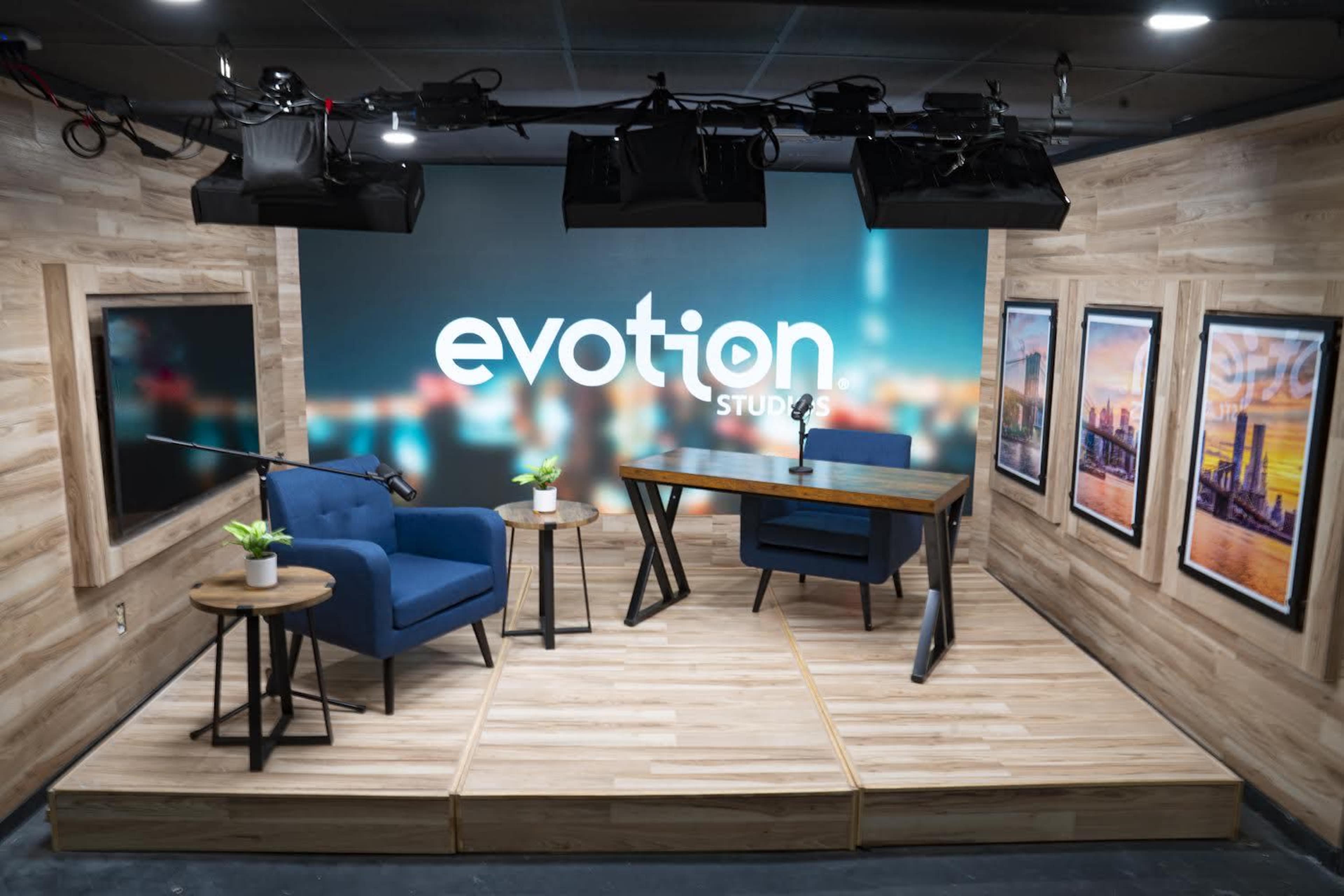 A modern studio setup features two blue chairs, a wooden table, and framed cityscape art on a wall with a large "evotion studios" logo.