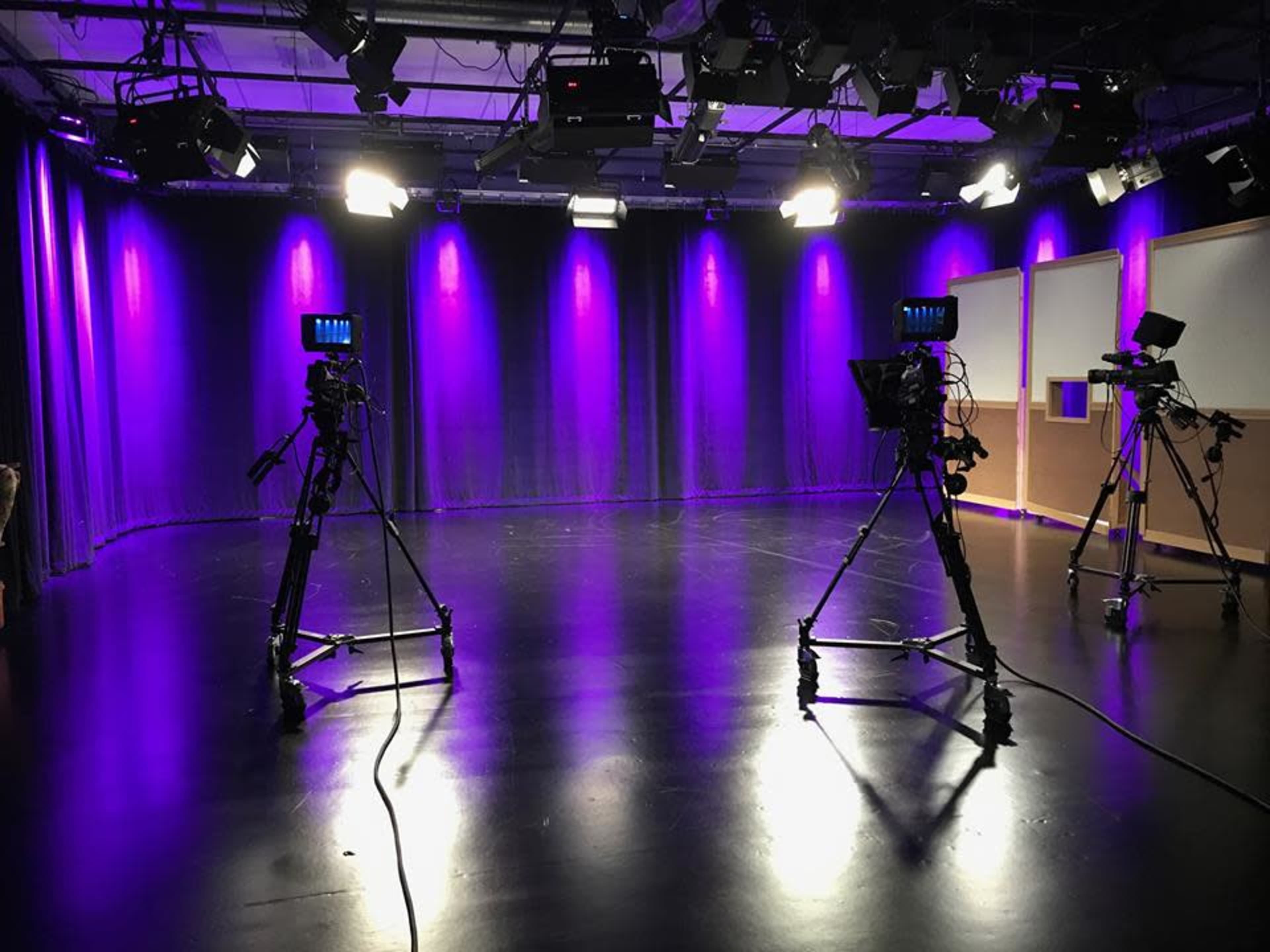 A film studio with two camera tripods positioned on a polished black floor, illuminated by purple stage lights.