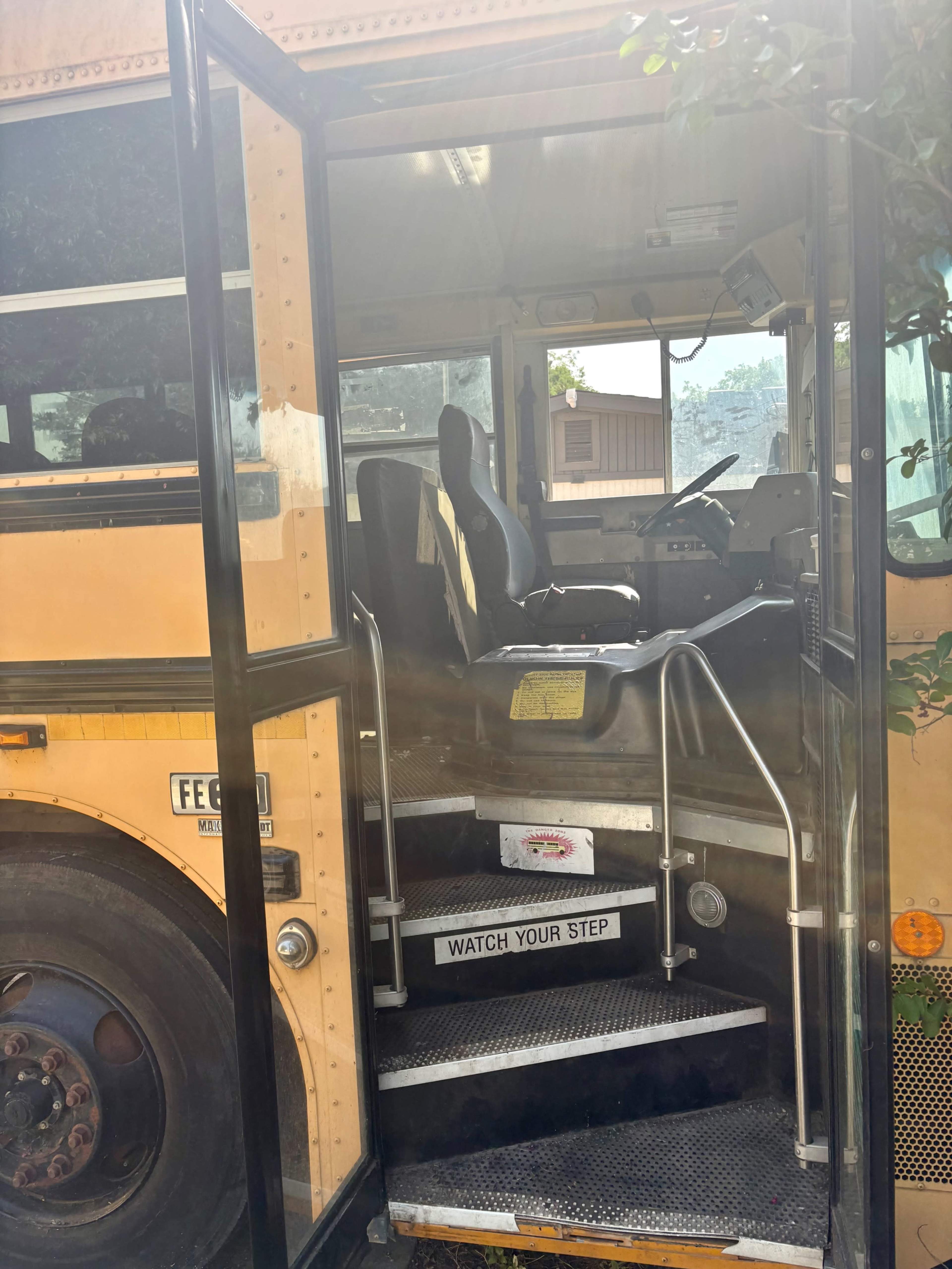 The image shows the entrance and driver's seat of a yellow school bus, with steps leading to the door and a sign that reads "WATCH YOUR STEP."