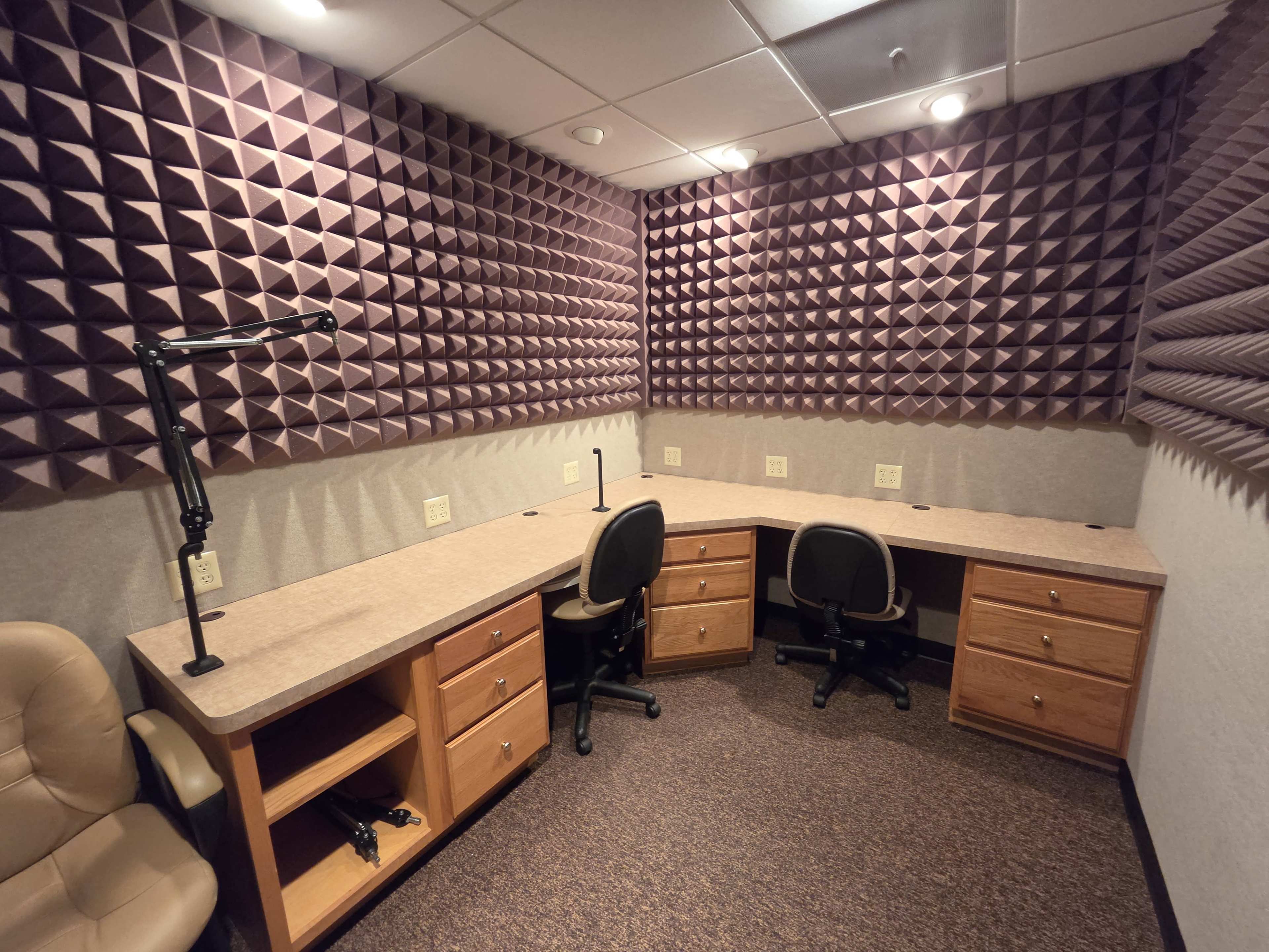 The image shows a soundproof recording studio with padded walls, a corner desk with two office chairs, and task lighting.