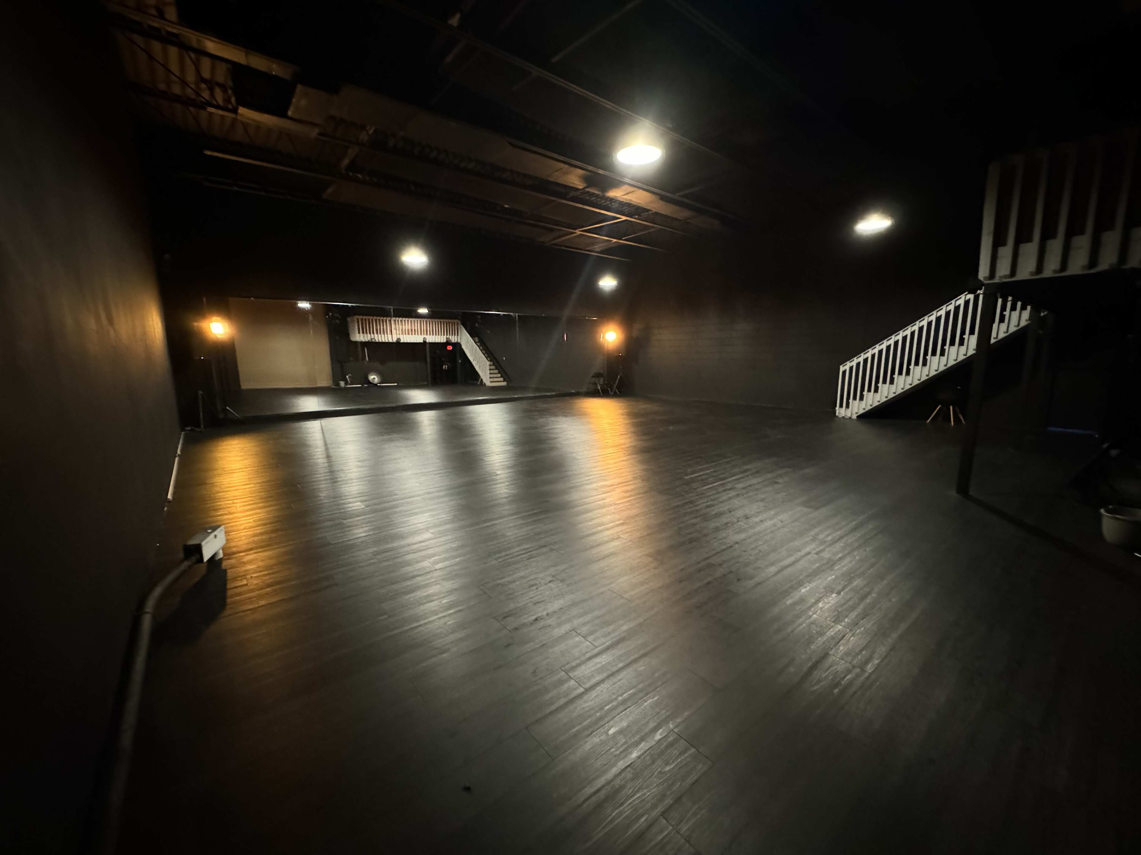 A spacious, dimly lit performance stage features dark wooden flooring and a set of stairs on one side.
